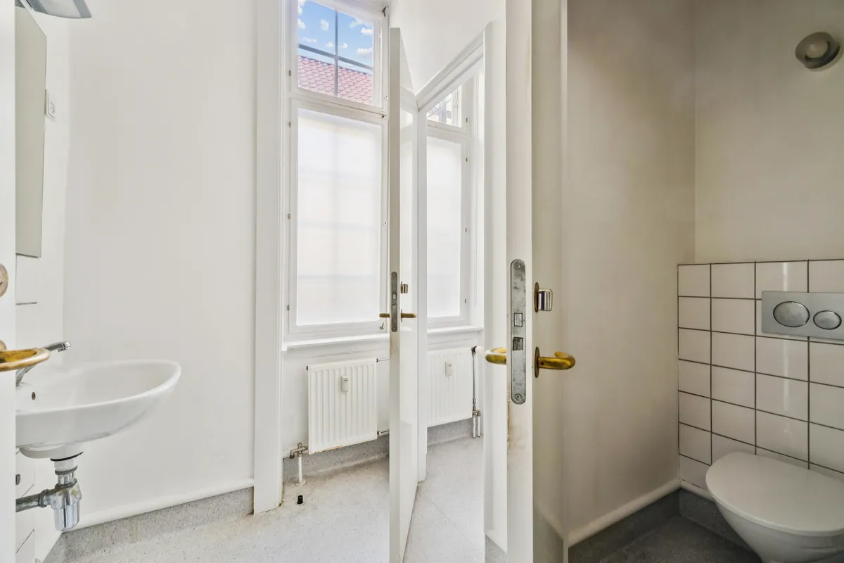 Foto af et rent og moderne badeværelse med en hvid toilet, hvid sænke med en guldfarvet vandhane og et vindue med en udsigt til Rådhusstræde.