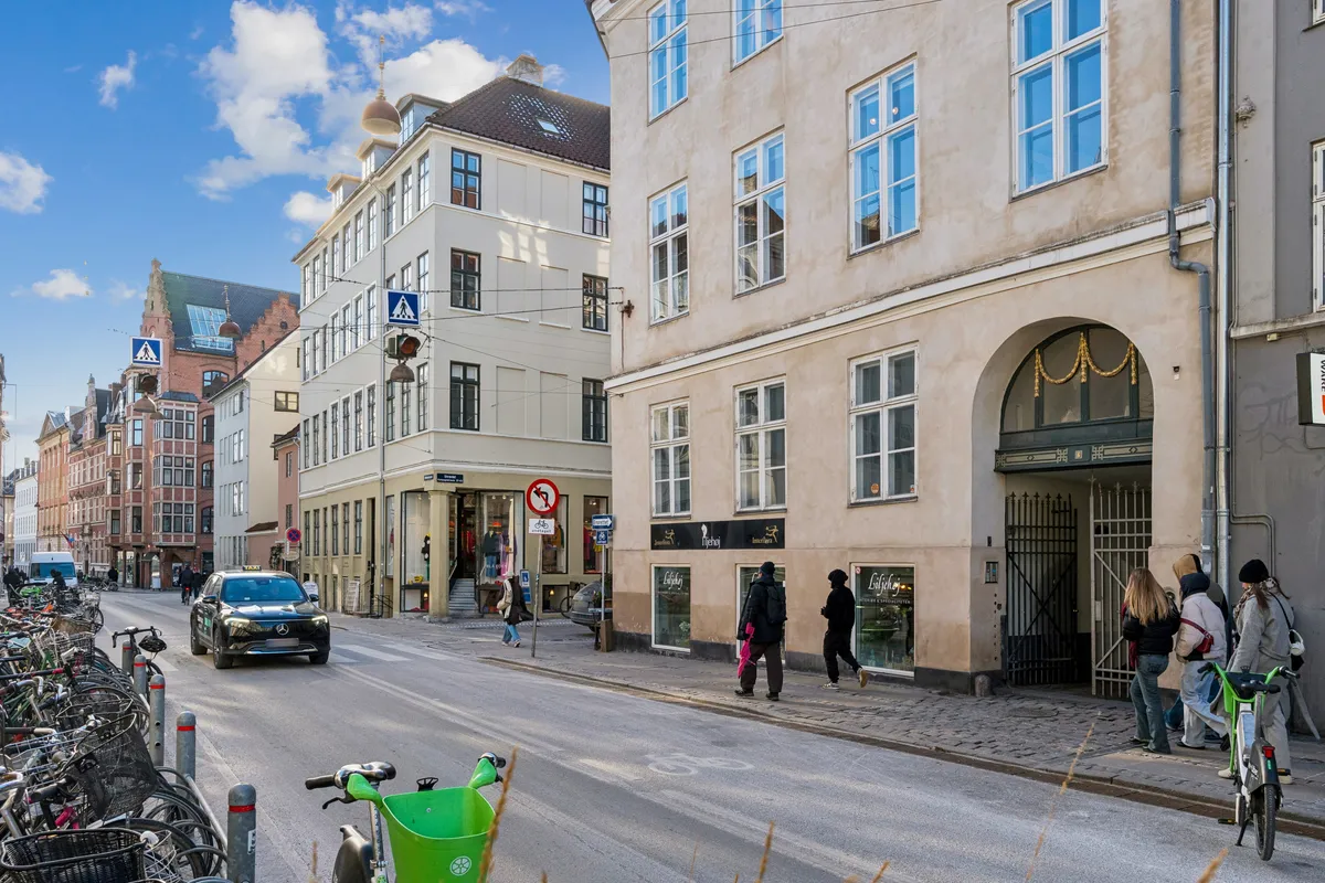 Mennesker og biler på Rådhusstræde og Kompagnistræde. En person med en grøn cykel og en med en hvid cykel. En Mercedes-Benz kører langs gaden. Mange cykler er parkeret på højre side af gaden.