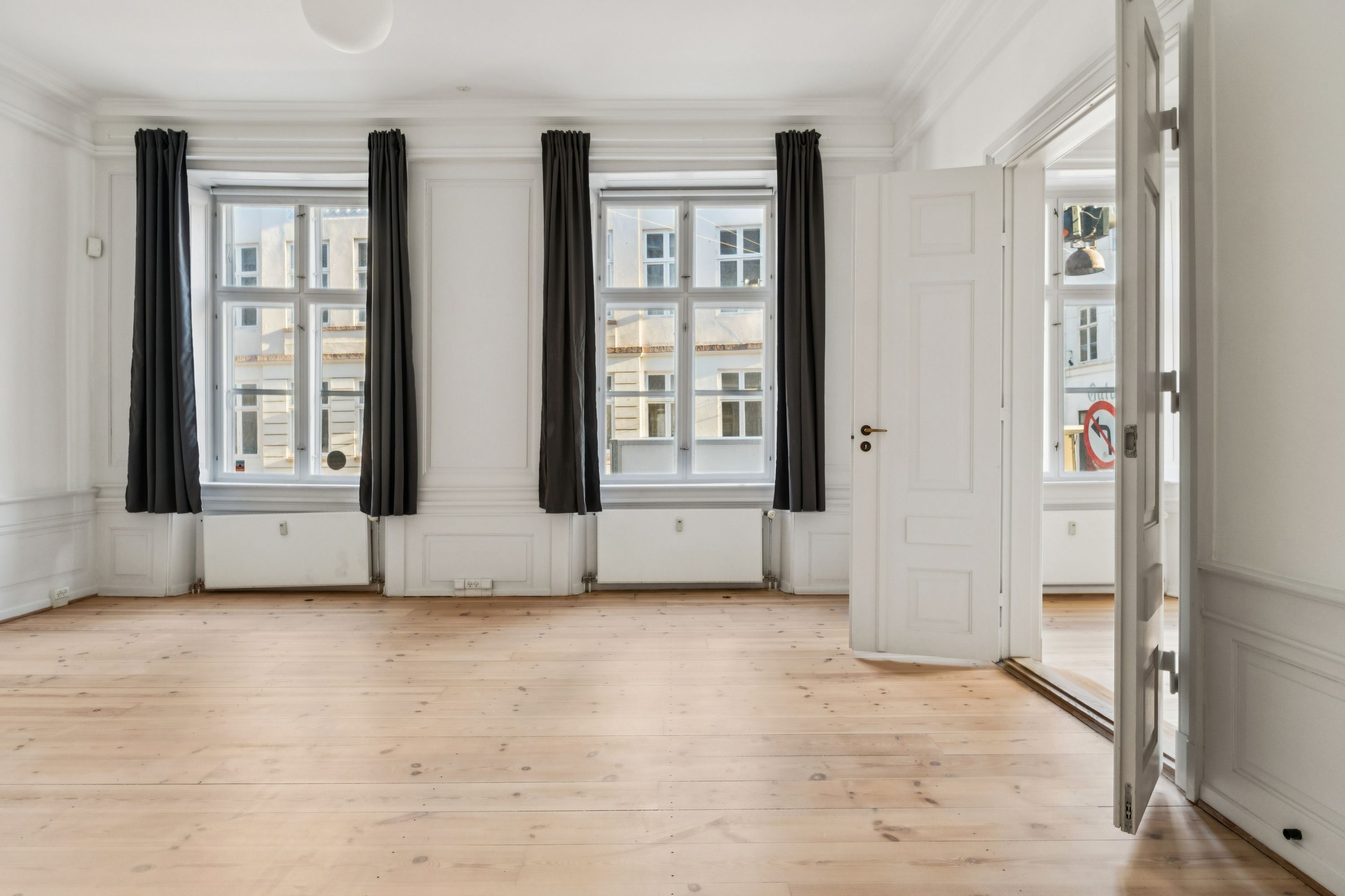 Lys, tom kontor med hårdt trægulv, hvide vægge og højt til loftet, to store vinduer med mørke gardiner og to hvide døre, der fører til det næste værelse
