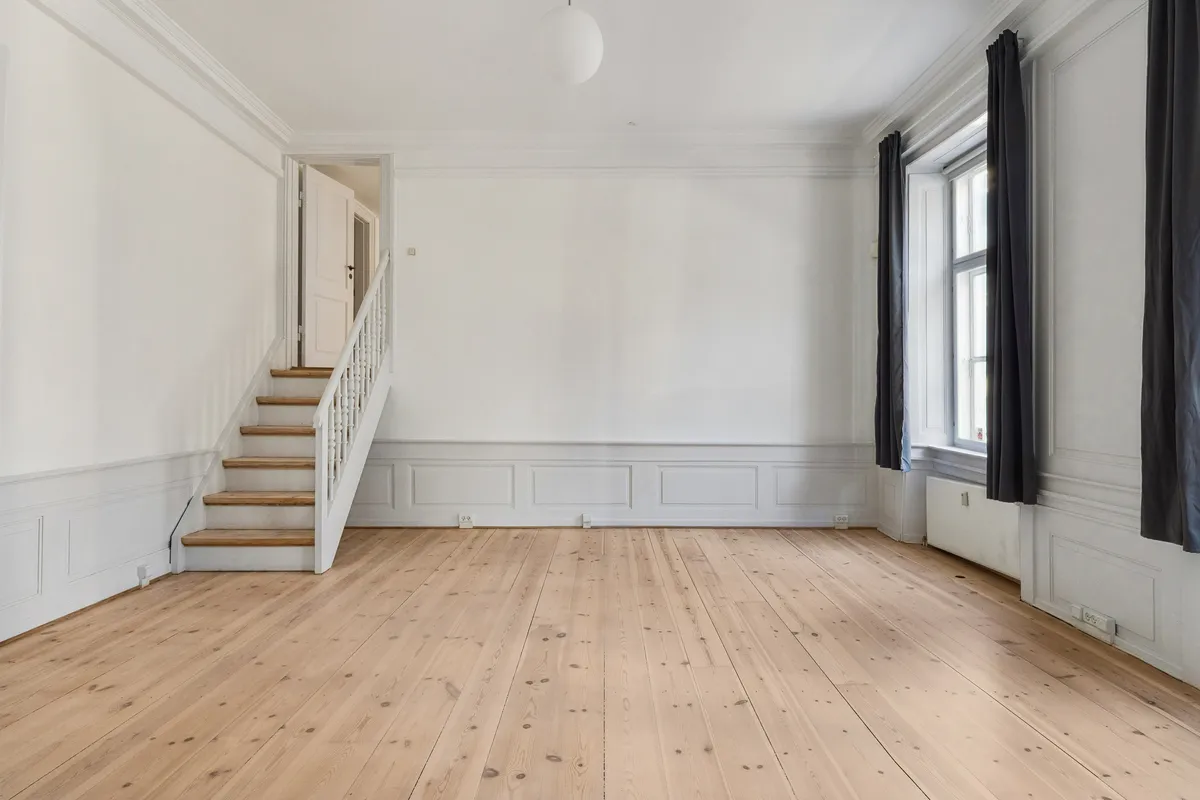 Et luksuriøst kontor med lyse trægulve og hvide vægge, hvor lysekronen hænger fra loftet og vinduet er dækket med mørke gardiner. Trappen er placeret på venstre side og fører op til næste etage.