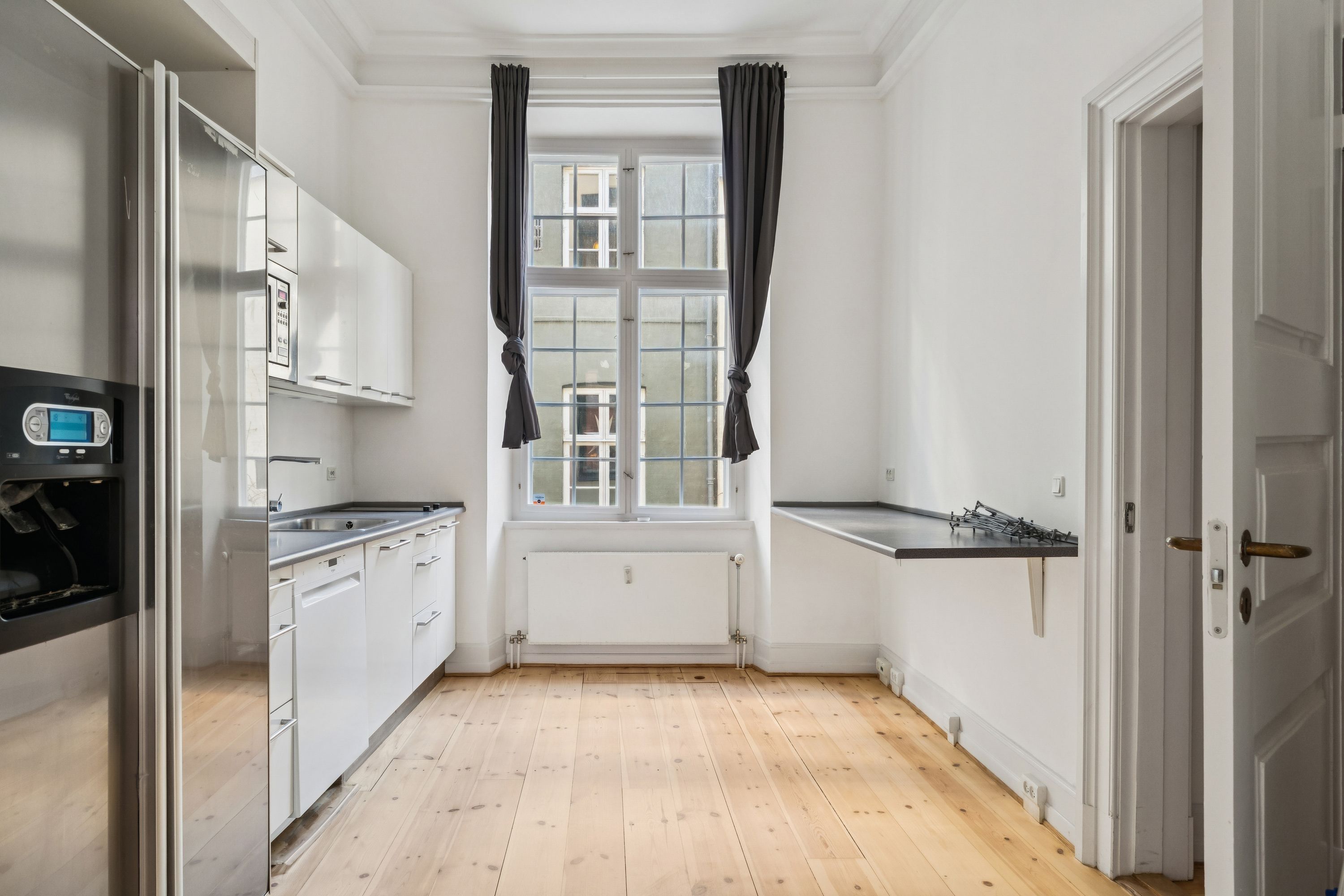 Et stort køkken med hvide vægge, lyse trægulve, en indbygget komfur, en stor køleskab og en hængende skorsten. Et vindue med dobbeltruder og gardiner i sort.