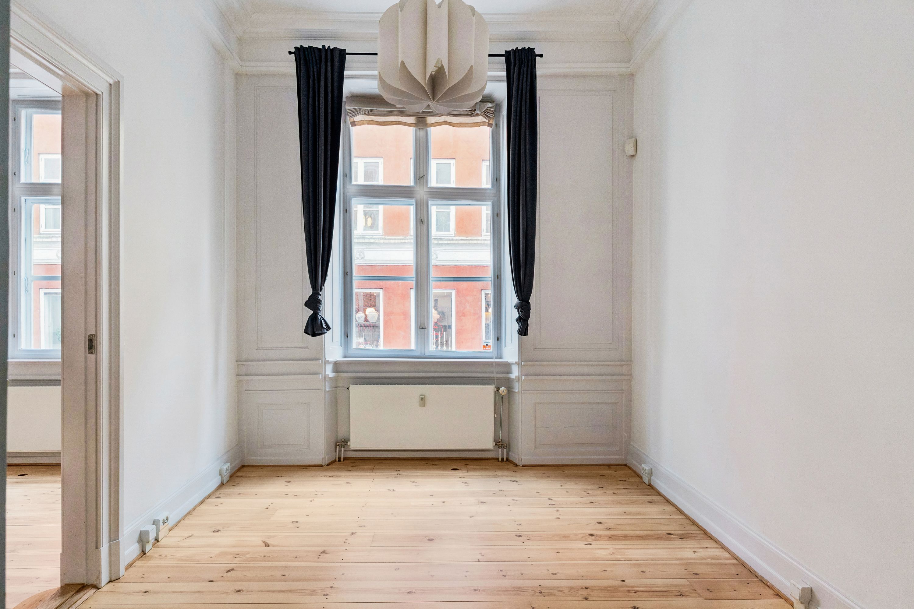 Lukket stue med lyse trægulve, hvidt malede vægge, to store vinduer med mørke gardiner og højt hængende lysekroner, radiator og døre mod åbne rum