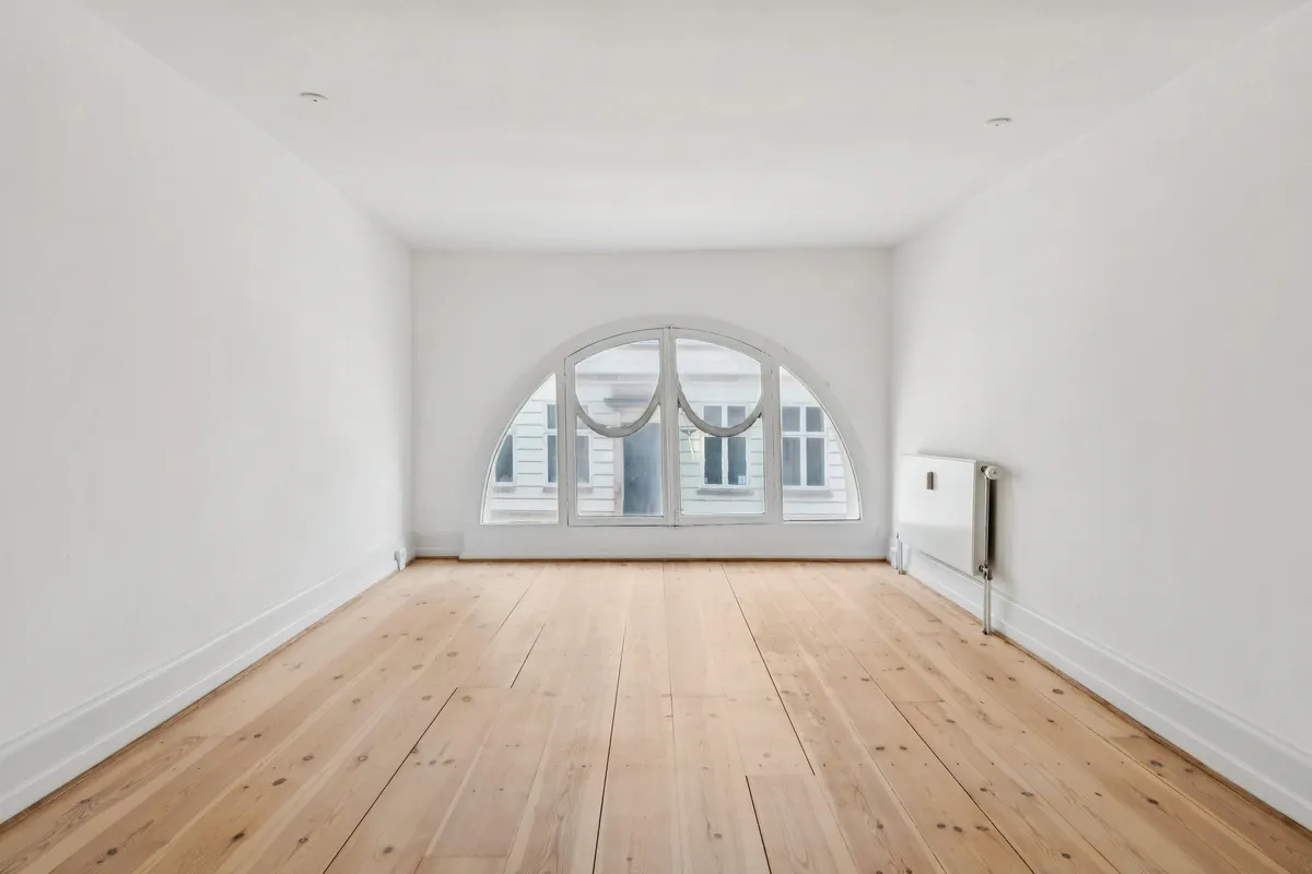 En tom kontorstue med hvidt loft og vægge, hårdt trægulv og en stor bueformet vinduesglas. Et radiator er placeret mod højre væg. Et hvidt hus med flere vinduer er synligt udenfor.