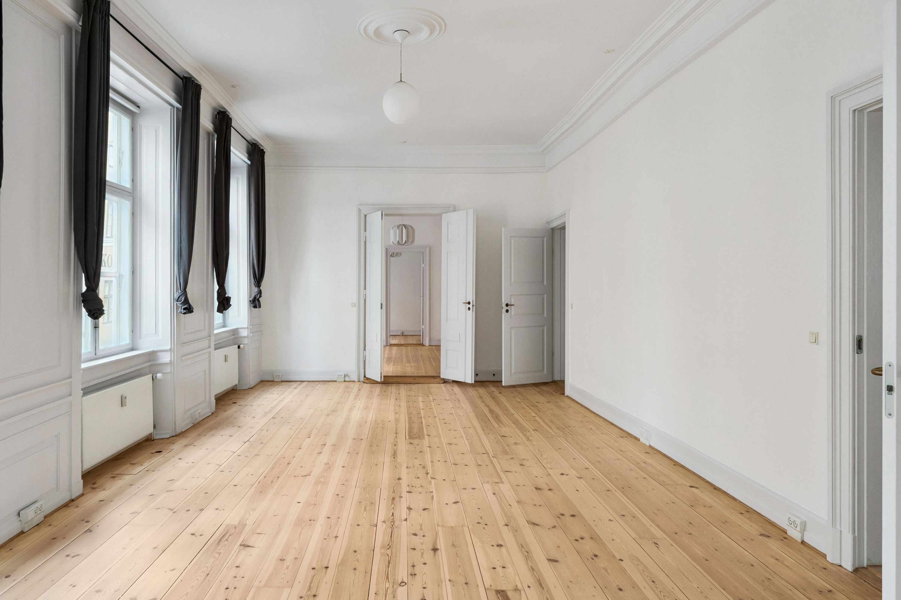 Lys kontor med hårdt trægulv og hvide vægge, store vinduer med sort gardin og åbne hvide døre til flere rum i baggrunden