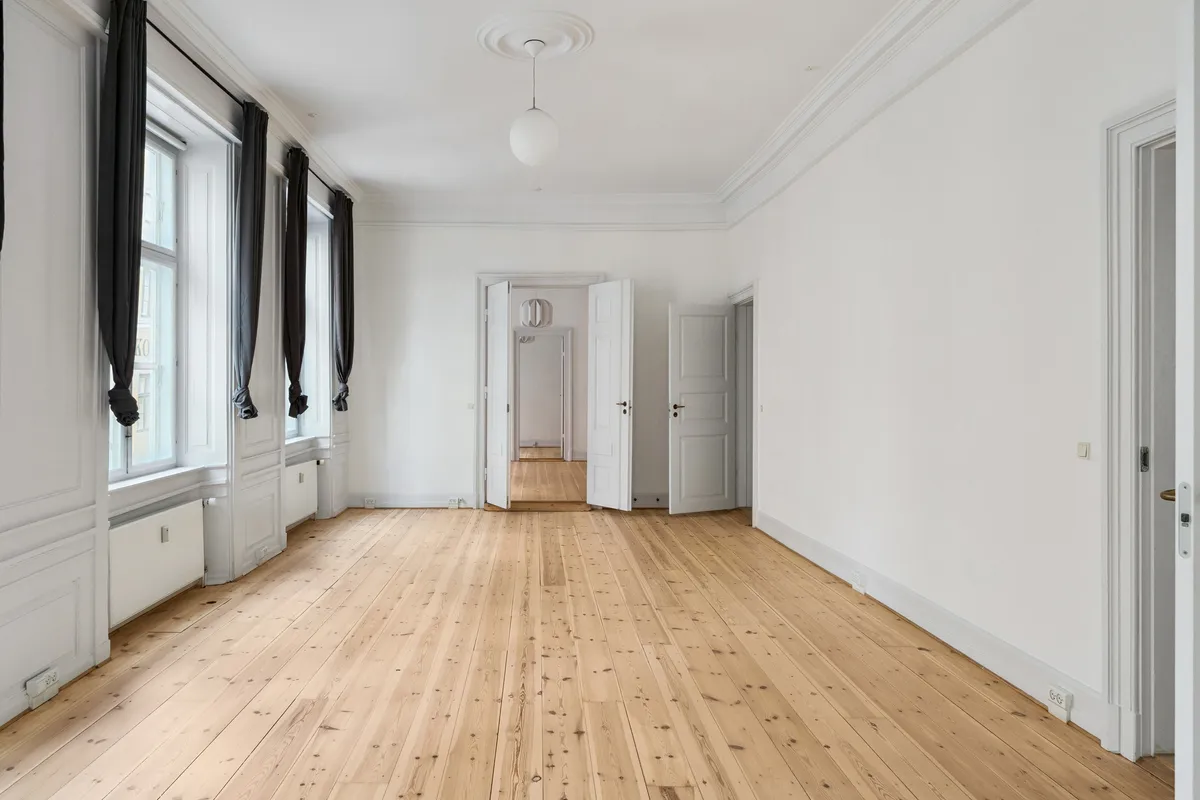 Lys kontor med hårdt trægulv og hvide vægge, store vinduer med sort gardin og åbne hvide døre til flere rum i baggrunden