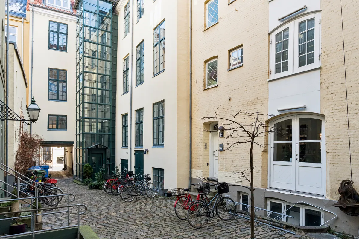 Kontor til leje i et slankt og stilfuldt bygningskomplekset med flere cykler parkeret udenfor. Bygningen har hvide facader og store vinduer. Et slankt træ står foran bygningen og der er en håndtag til cykelhænge.