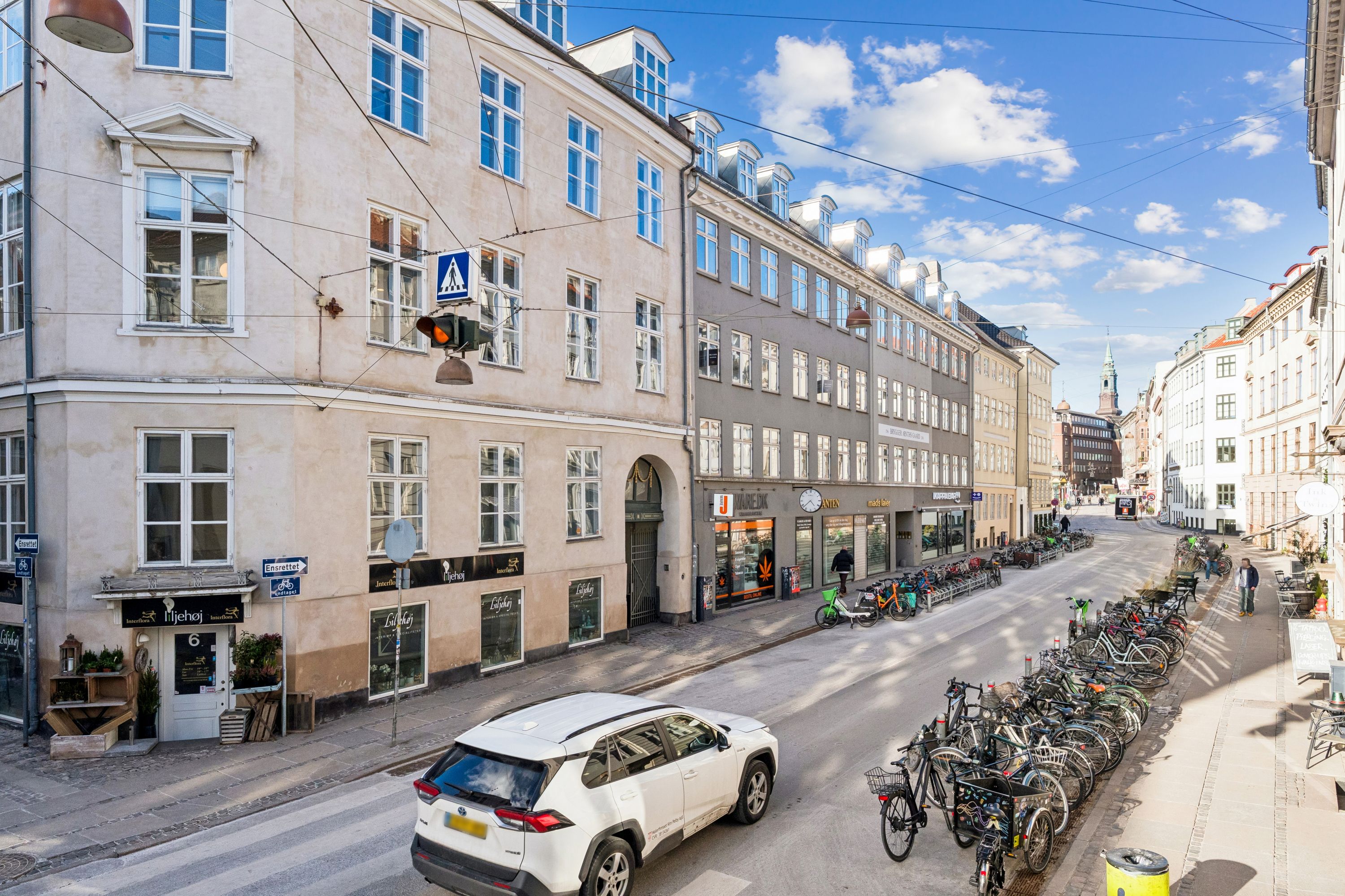 En hvid SUV kører langs en gade med parkerede cykler, forretninger og bygninger i baggrunden, under en blå himmel med skyer