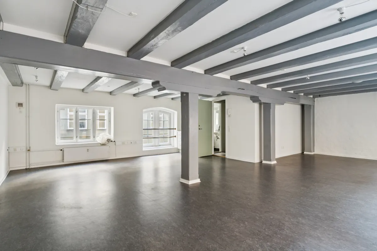 Et tomt kontor med lyse vægge, slanke søjler og lysegrå træbjælker i loftet. Store vinduer og en radiator under vindueskarme.