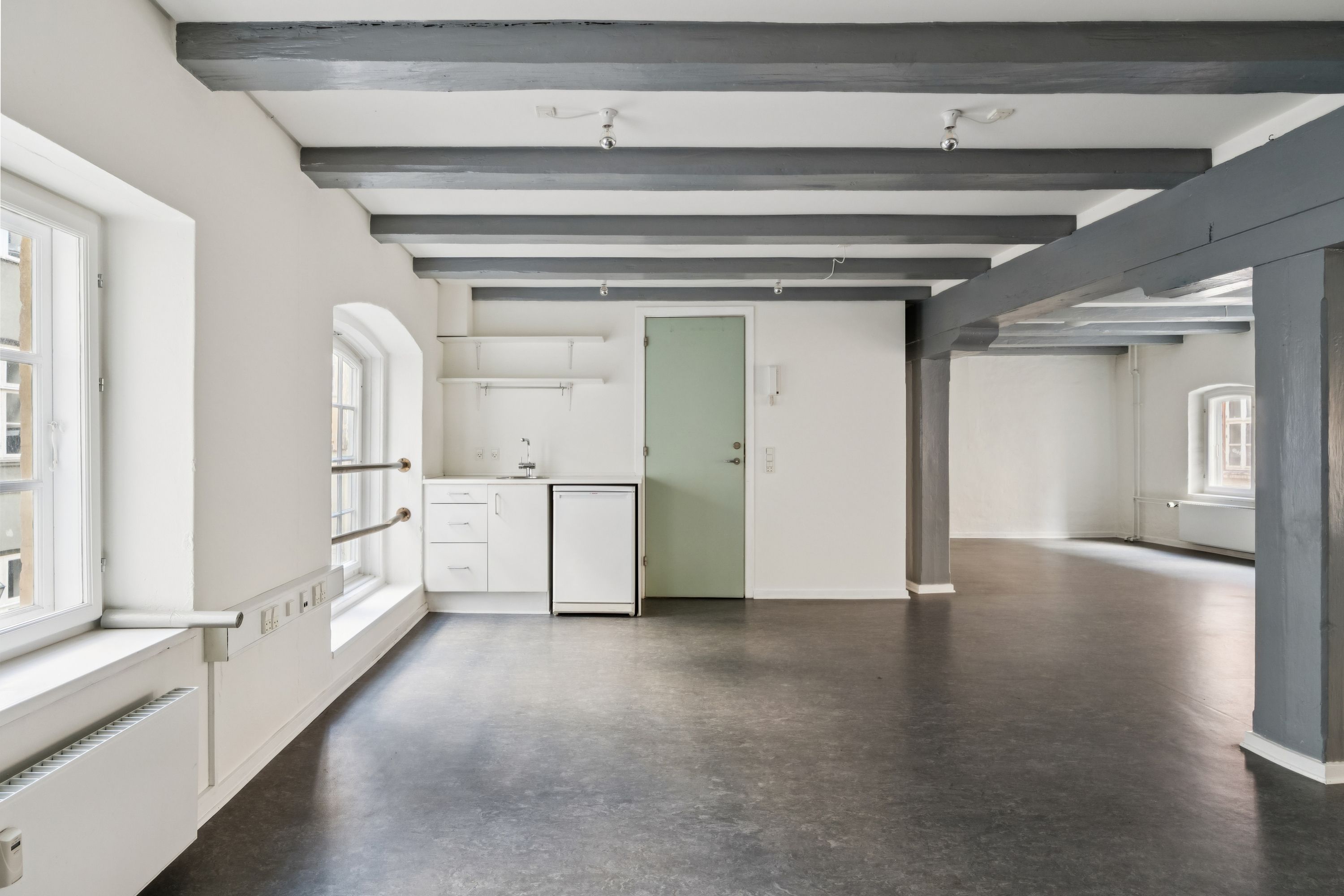 Kontor med højt loft, hvide vægge, lysegult lys, møbler og lysekroner i loftet, og et køkken med hvide vægge og en grøn dør