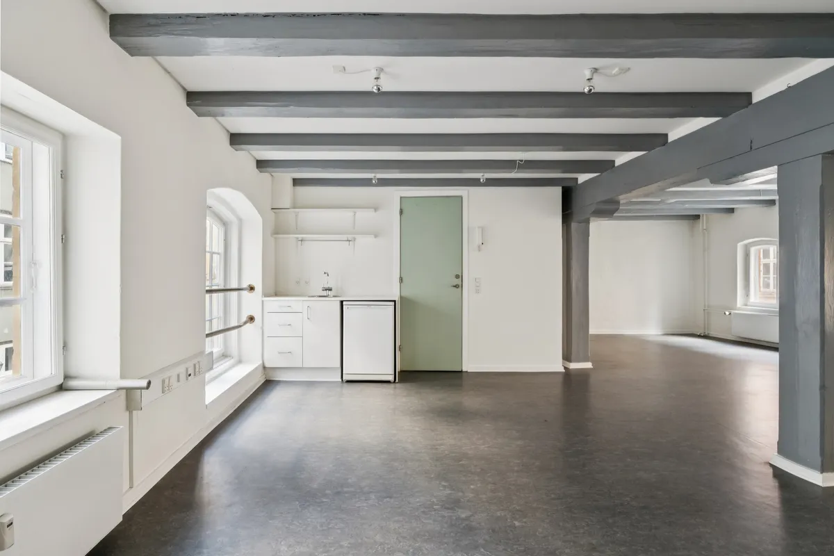 Kontor med højt loft, hvide vægge, lysegult lys, møbler og lysekroner i loftet, og et køkken med hvide vægge og en grøn dør