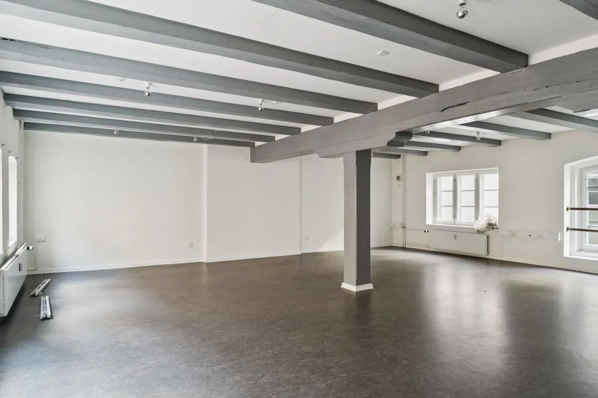 Et stort, tomt rum med hvide vægge, lysegrå gulvbelægning, lysegrå bjælkeloft og lysegrå søjler med hvide sokkelplader