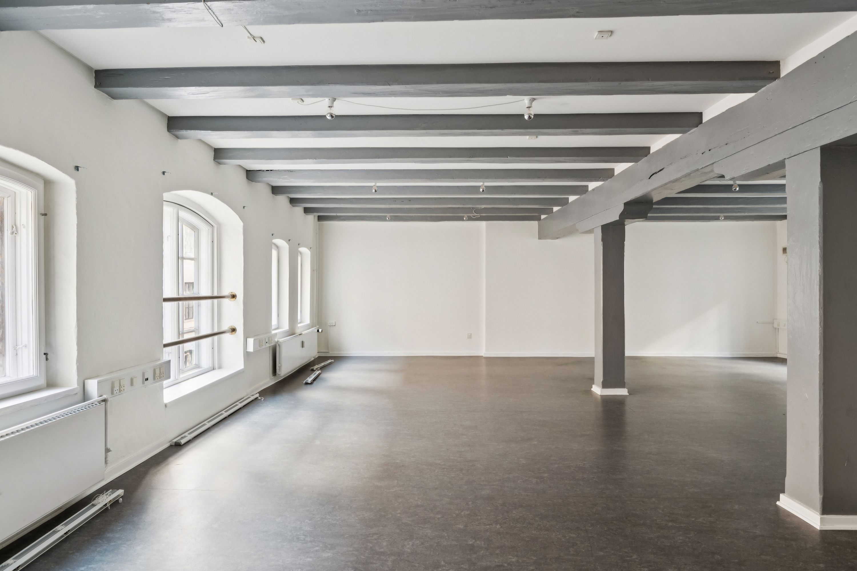 Moderne kontor i 1. sal med højloft, lyse vinduer og stilfulde bjælker. Minimalistisk design med hvid væg og grå gulv. På vindueskarme står radiatorer, der forsyner lokalet med varme.