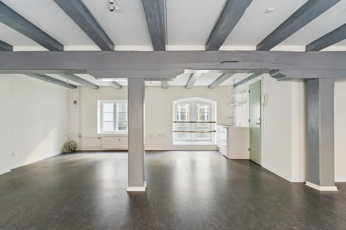 Værelse med højt loft, gulv og vægge, to vinduer, hvidt køkken med en smal hylde og et hængende telefonapparat, lysegrå bjælker, to lyse, hvide søjler og gulvbelægning