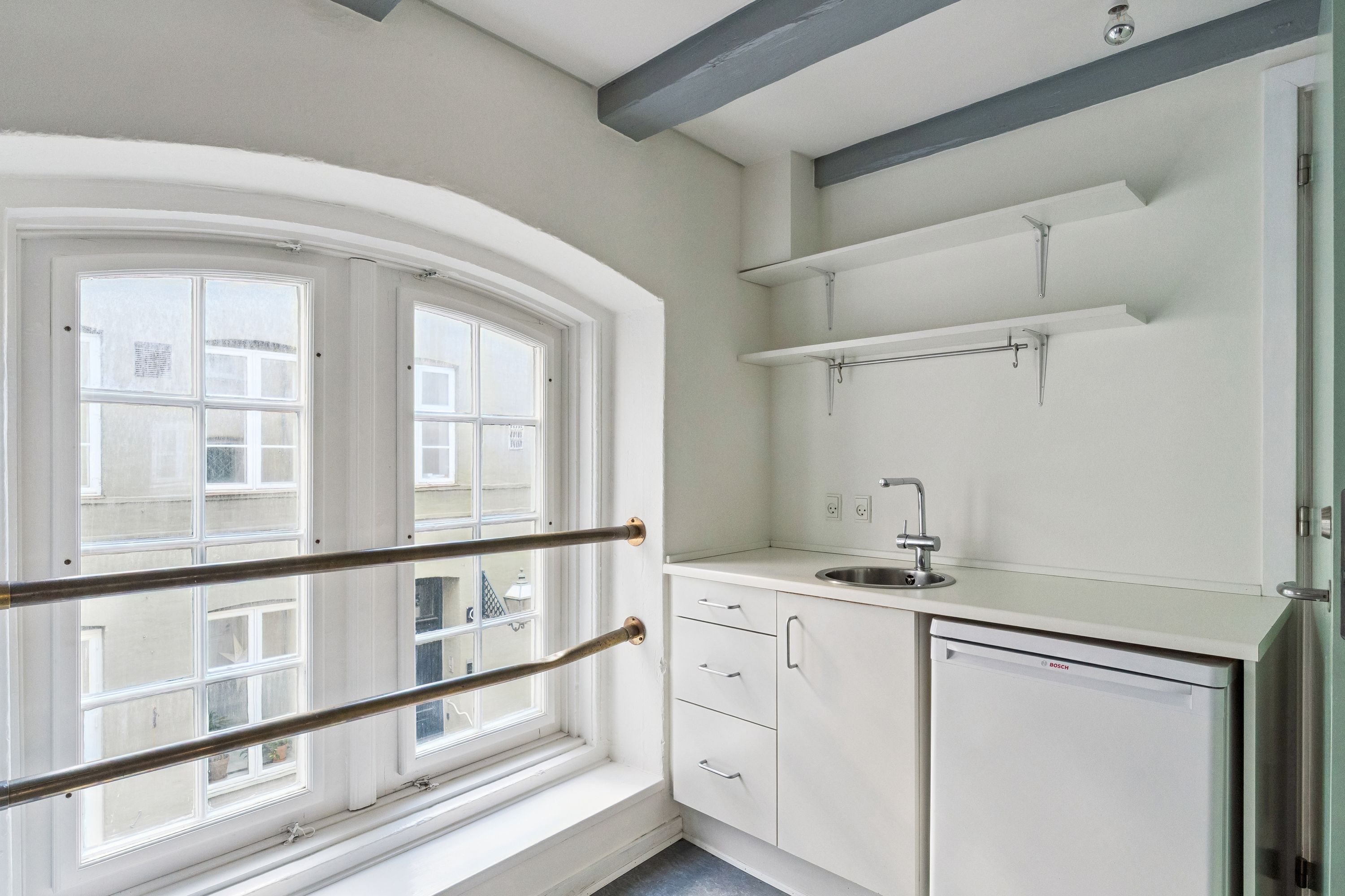 En lille køkken med hvide vægge, lysegule loftbjælker og et hjørne med vaskemaskine og vask under vindue med udsigt til Københavns gadebelysning