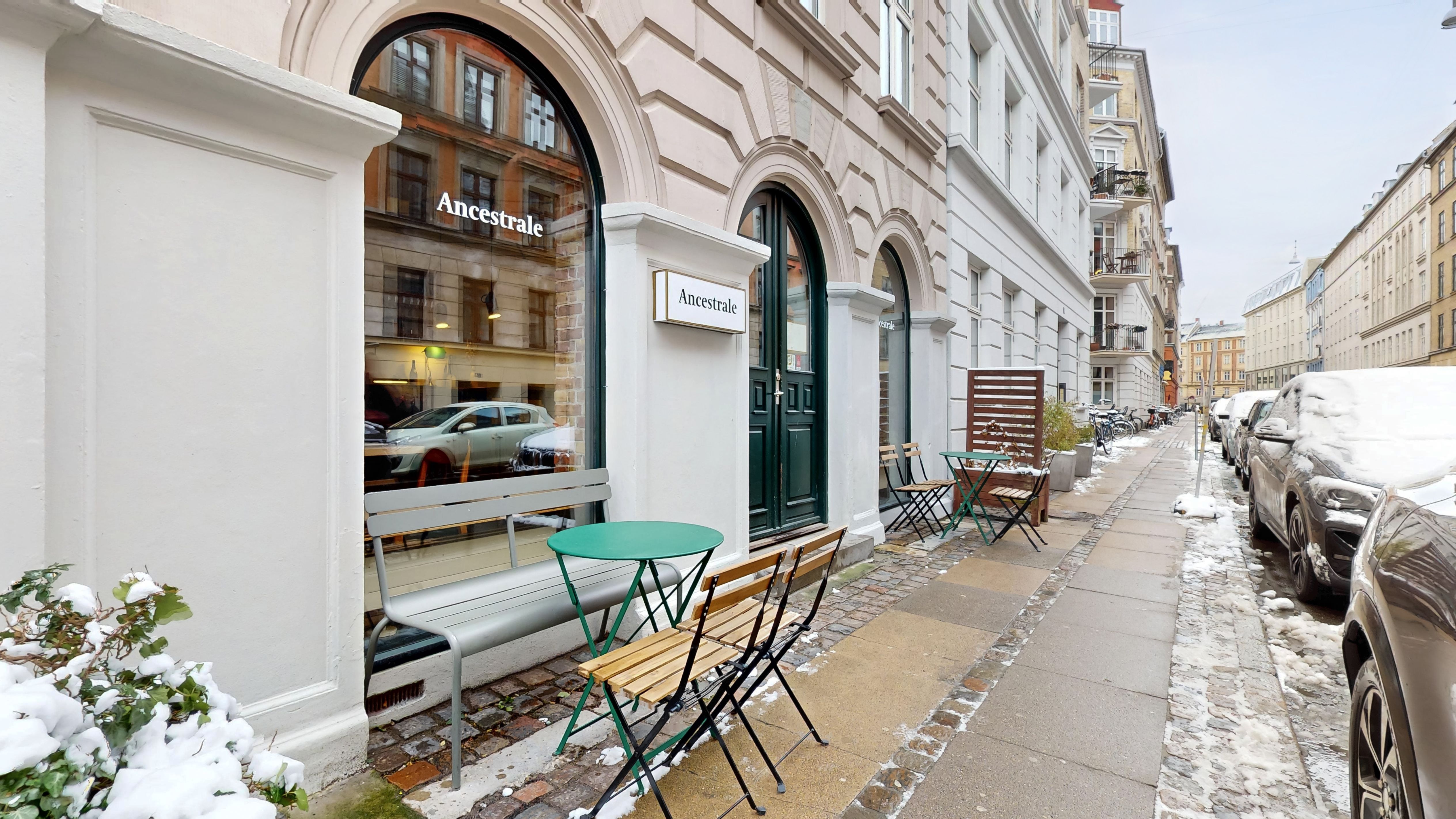 Hvid malet bygning med stolpe og udstillingsvindue med restaurantnavnet Ancestrale. To grønne stolpeborde med stole og en række parkerede biler i baggrunden.