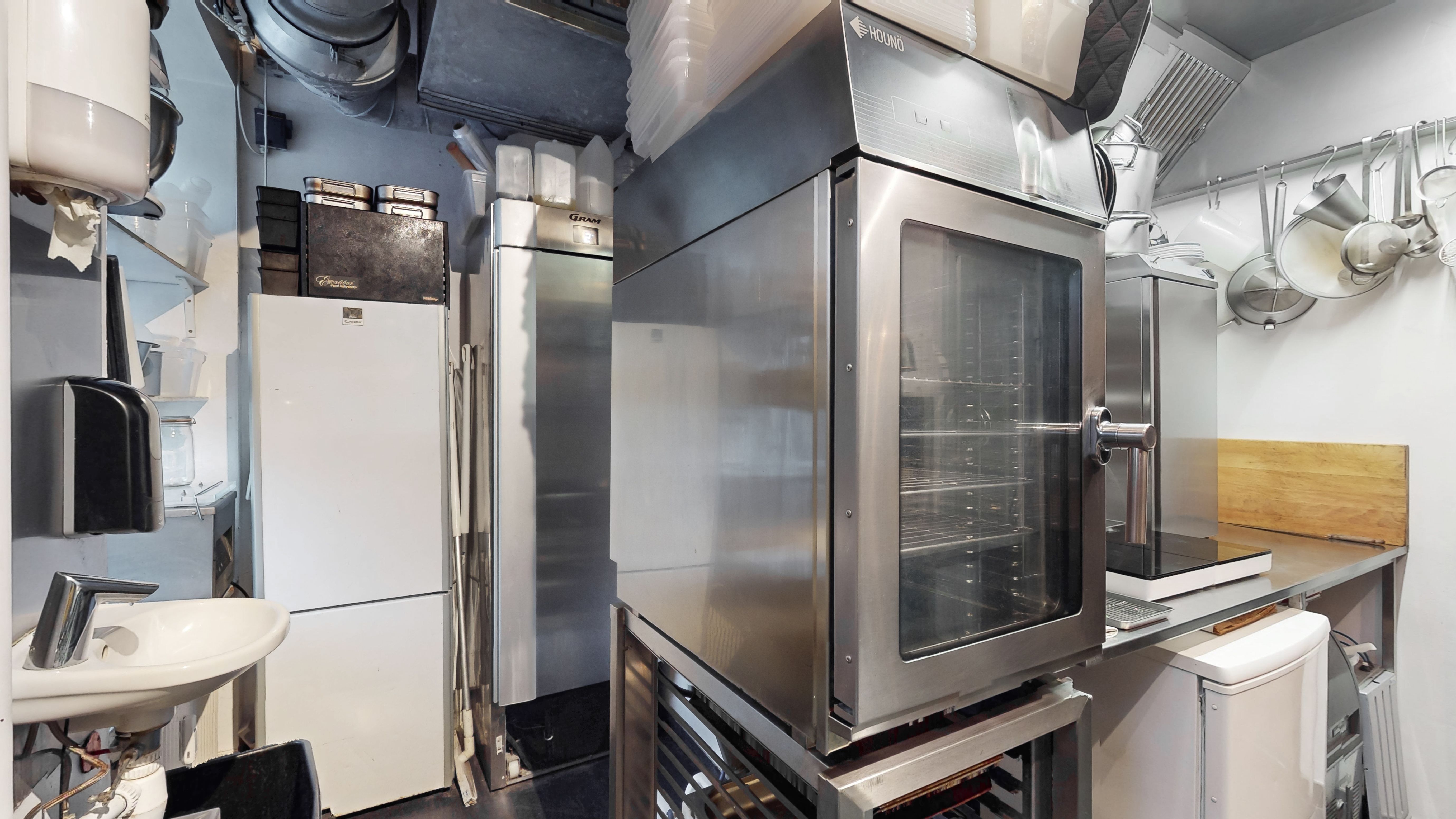 Professionelt køkken med rustfri ovn og køleskab, skraldespand, lyse køkkenhylder og stålhængsler. Vægge med opbevaringsbokse og hængende kogegrej.