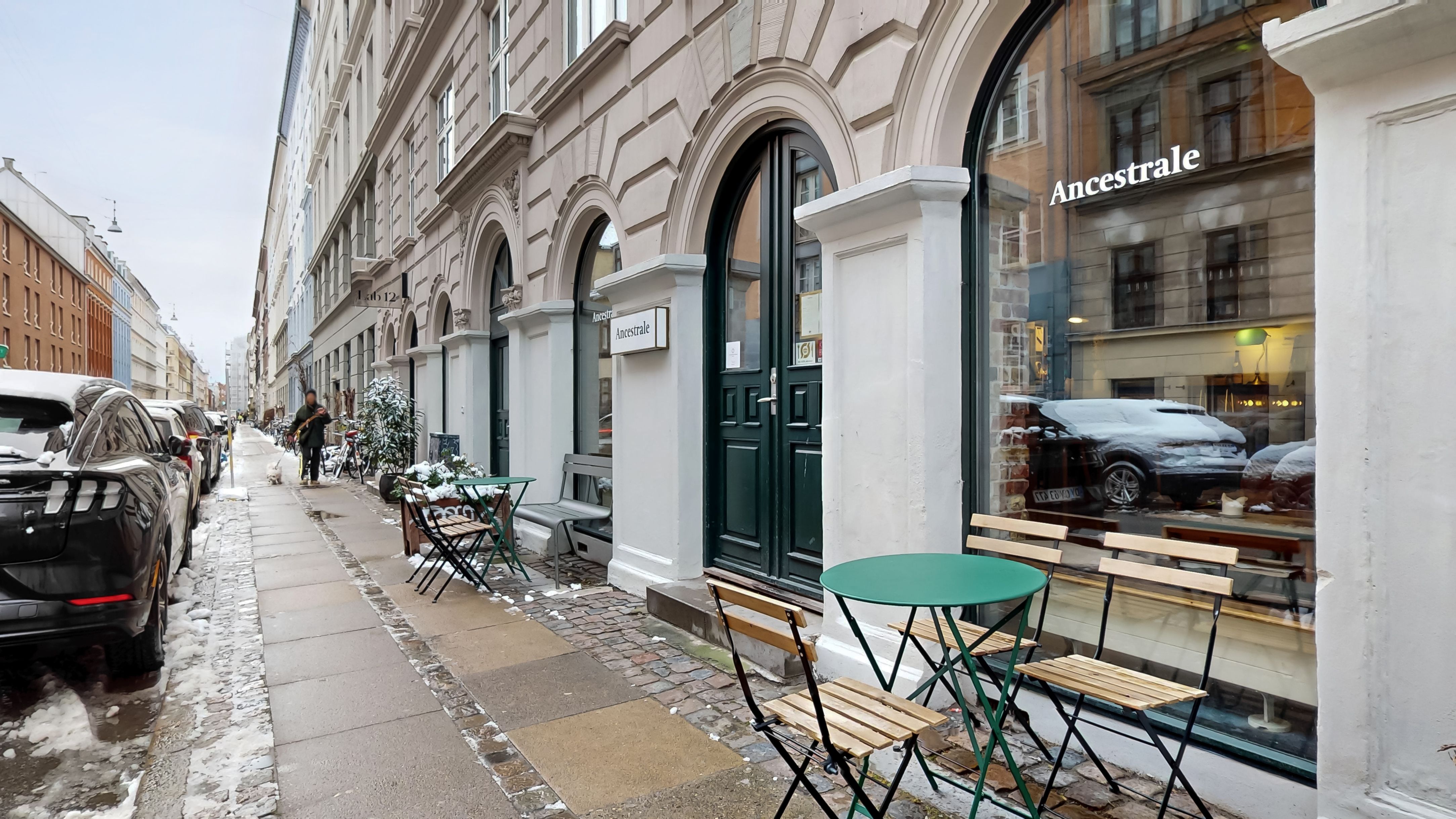 Lejlighed med restaurant på Oehlenschlægersgade 12. En lille grøn rund bord med stole uden for restauranten. Et par biler parkeret med sne. Og en person der går på fortovet.