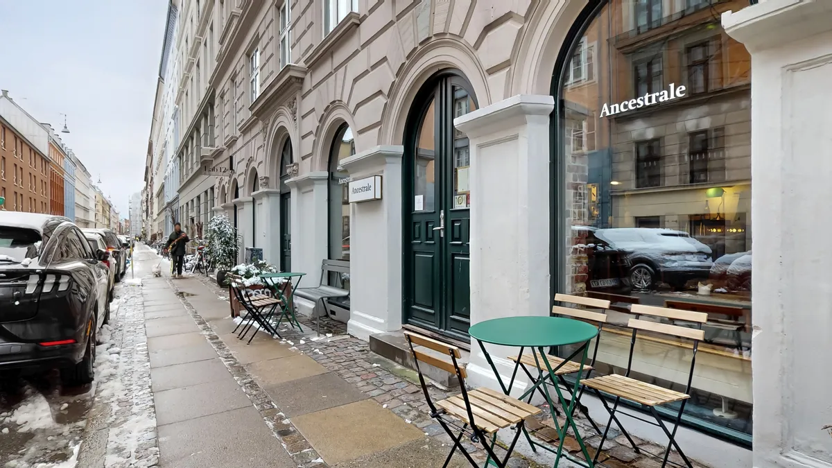 Lejlighed med restaurant på Oehlenschlægersgade 12. En lille grøn rund bord med stole uden for restauranten. Et par biler parkeret med sne. Og en person der går på fortovet.