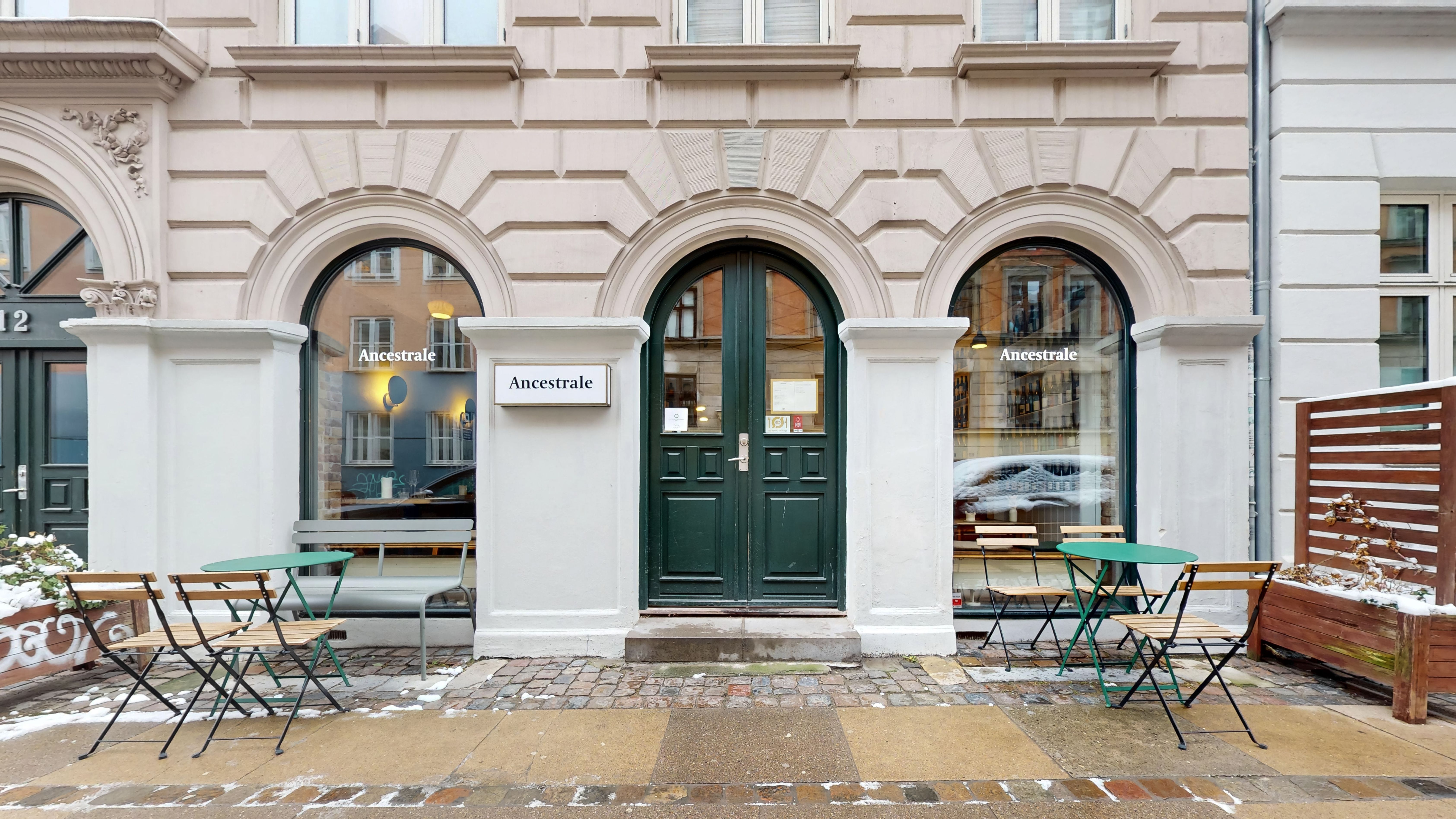 Valgmulighed for en restaurant til afståelse på Oehlenschlægersgade 12 i København. Opholdsområde med borde og stole. Vinduer og hoveddør med mærket "Ancestrale". Sne dekker gåbanen.