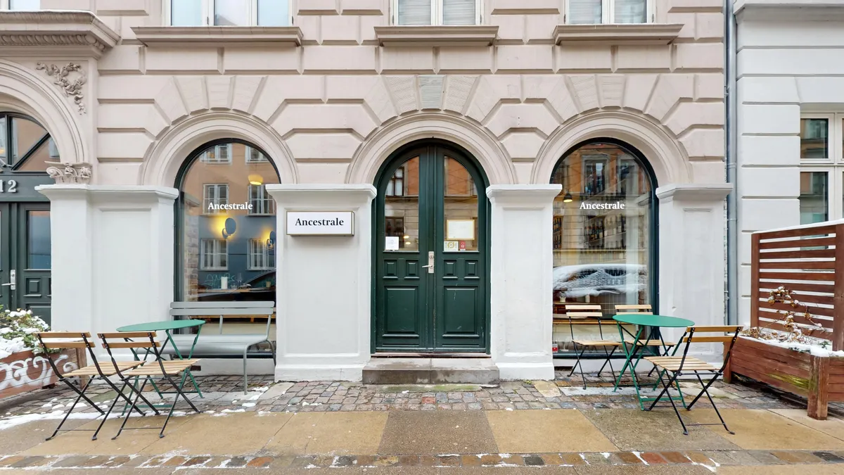 Valgmulighed for en restaurant til afståelse på Oehlenschlægersgade 12 i København. Opholdsområde med borde og stole. Vinduer og hoveddør med mærket "Ancestrale". Sne dekker gåbanen.