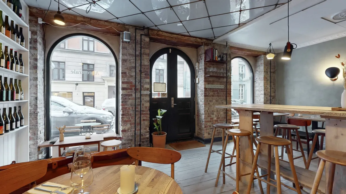 Restaurant med vinduer og stole. Køkken med borde og støtter og vinflasker på hylder. Lampe lyser og plante i potte.