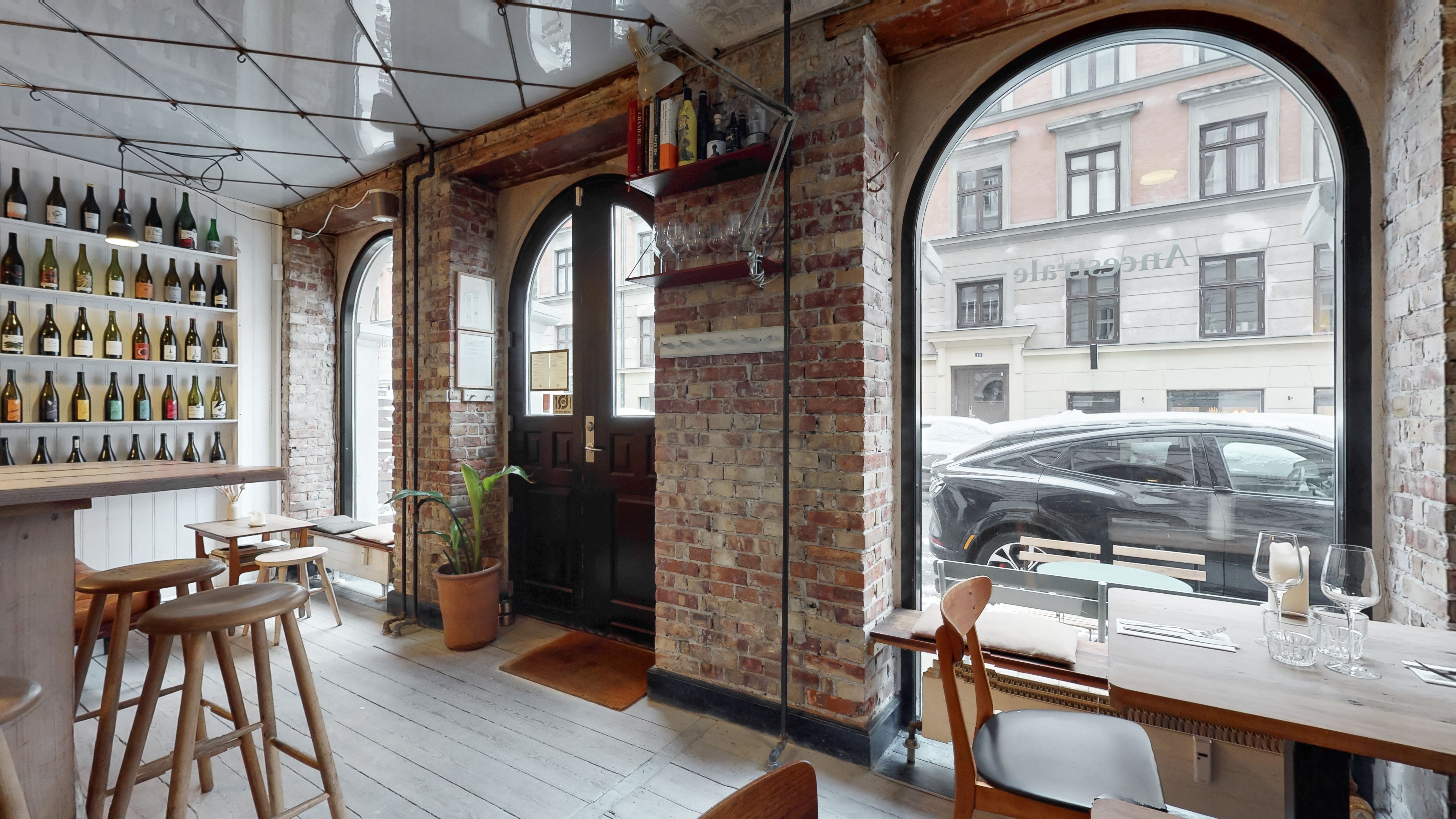 Restaurant med vinduer, træborde, stole, plante, vinflasker, og udsigt til biler og bygninger på Oehlenschlægersgade 12, 1663 København V.