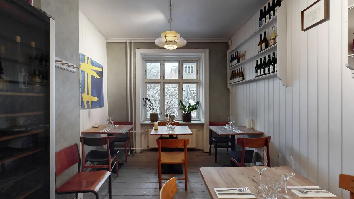 Interiør med to borde, stole, vinglas og en plante i vindueskarme. Dekorative kunstværker og et vinkøleskab med flasker.