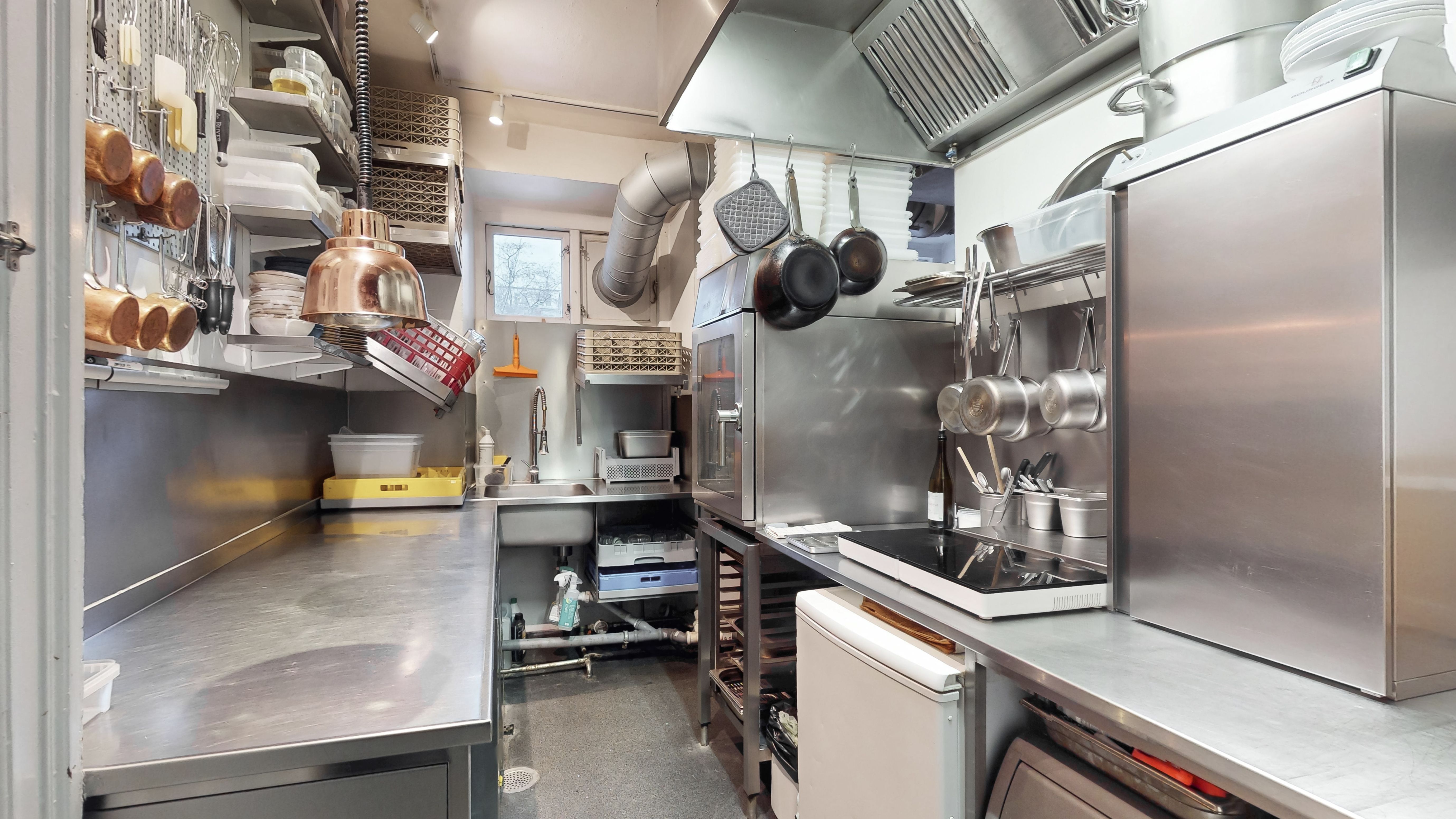 Moderne kommercielt køkken med flere apparater og udstyr samt lyse loftslampe. Væggen er forsynet med opbevaringssystemer og ståhylder.