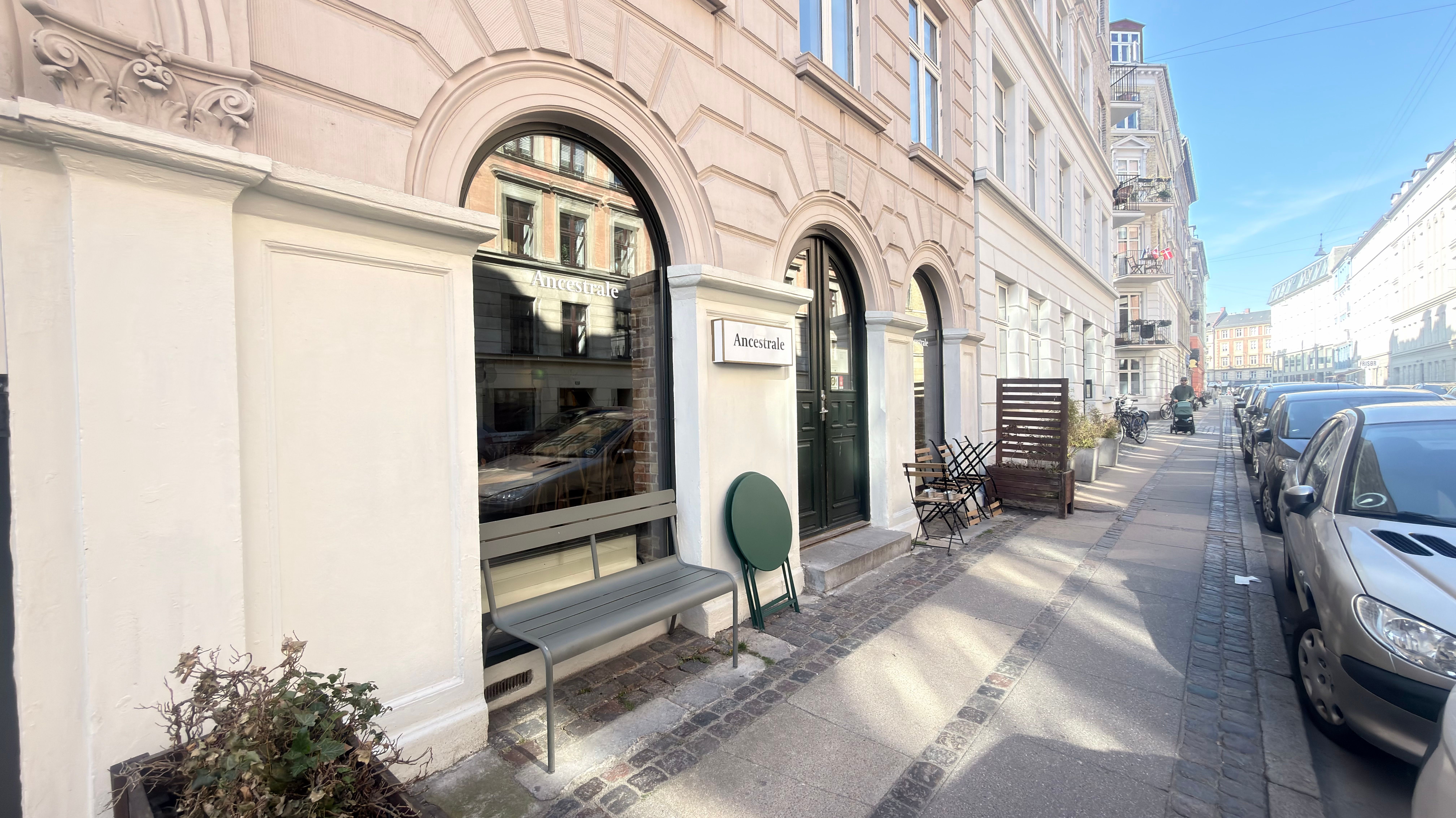 Grønne stole og bænke på gågaden med restauranten Ancestrale og biler parkeret på siden