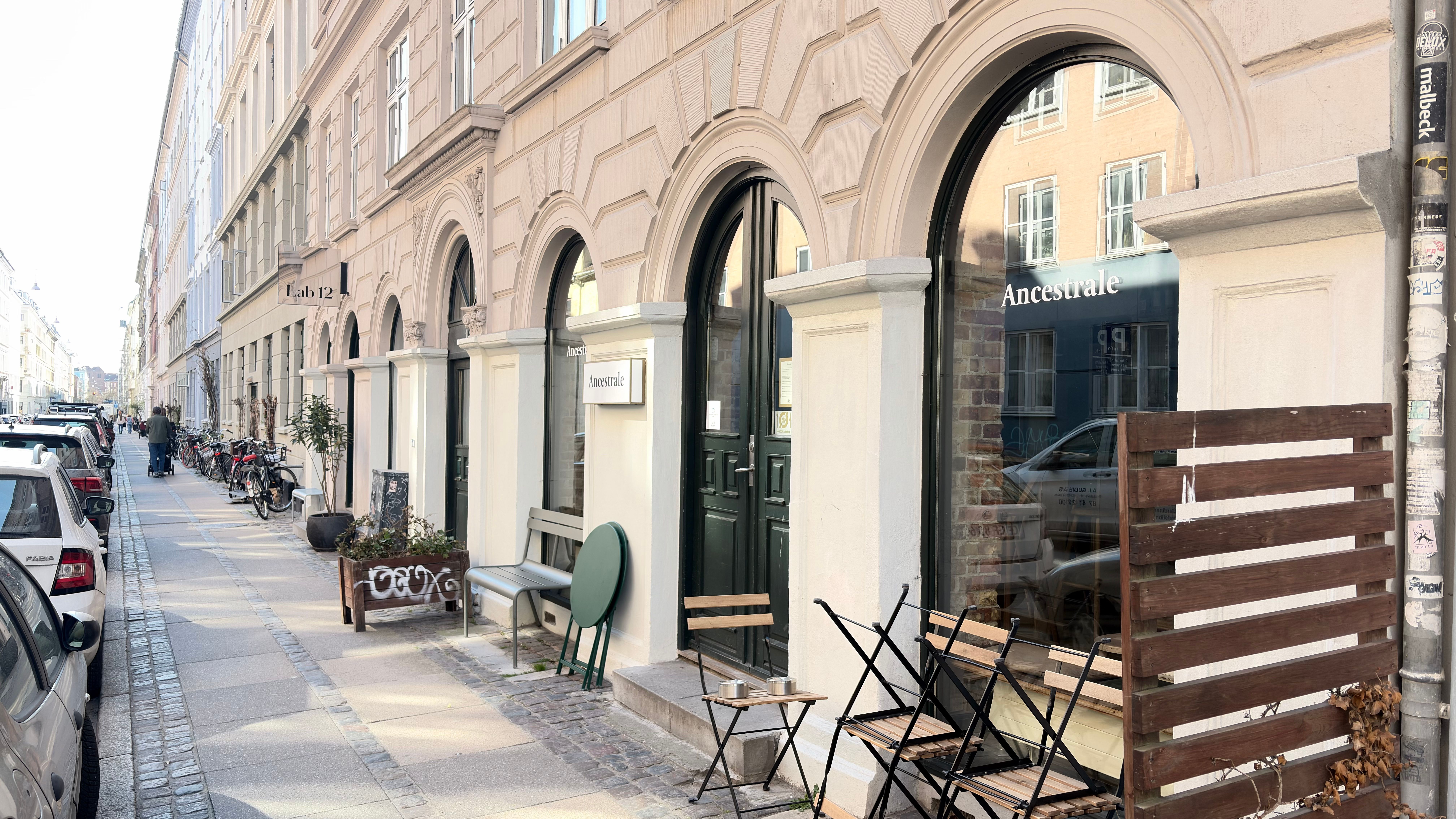 Restaurateuren Ancestrale med grønne stole og bord, hvor der er parkerede biler og cykler langs gaden