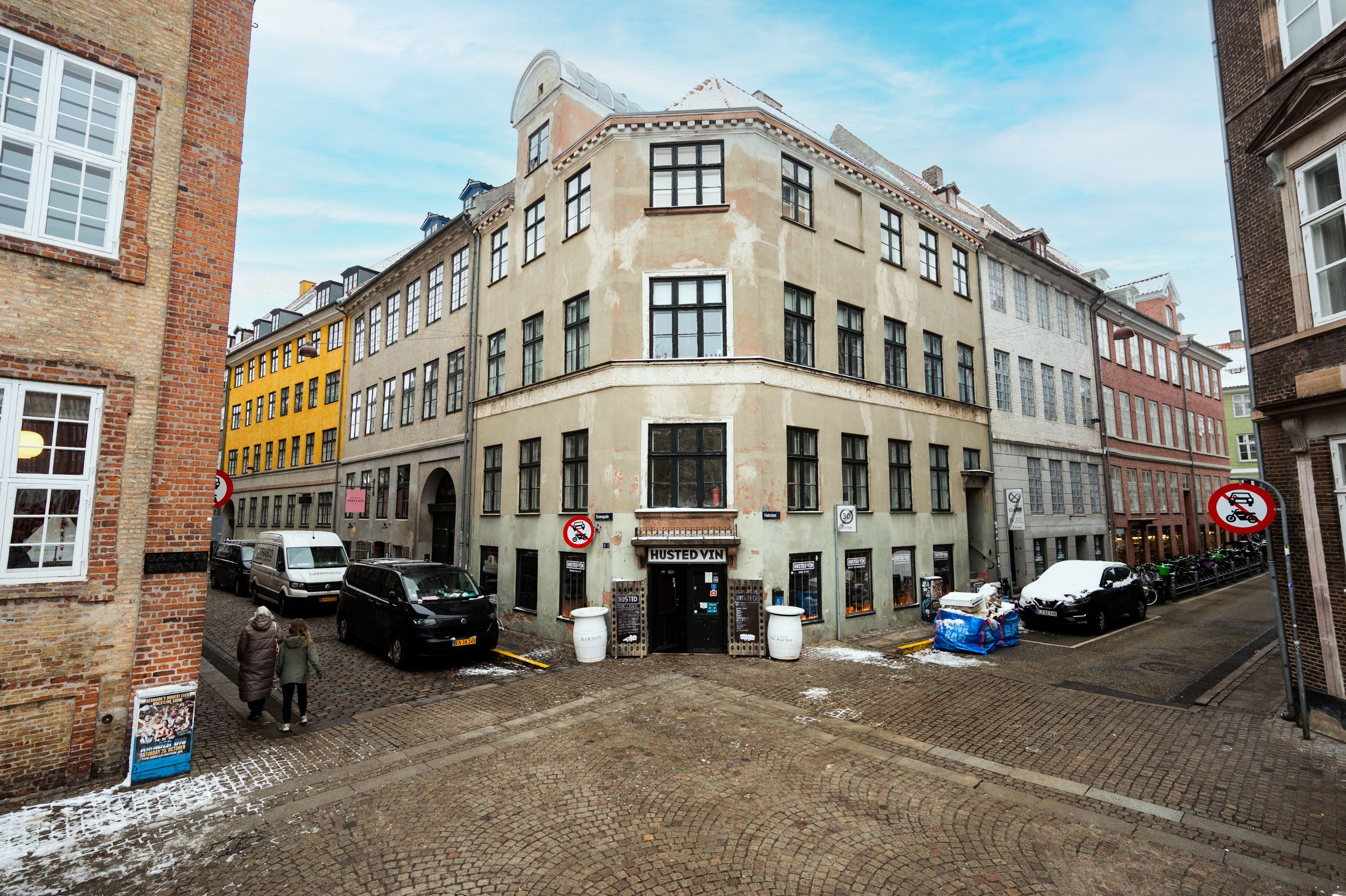 Købmandsgård fra 1700-tallet med historiske detaljer, hvor husfronten er beskidt og butikken hedder Husted Vin, og to personer går langs gaden med parkerede biler og søjler