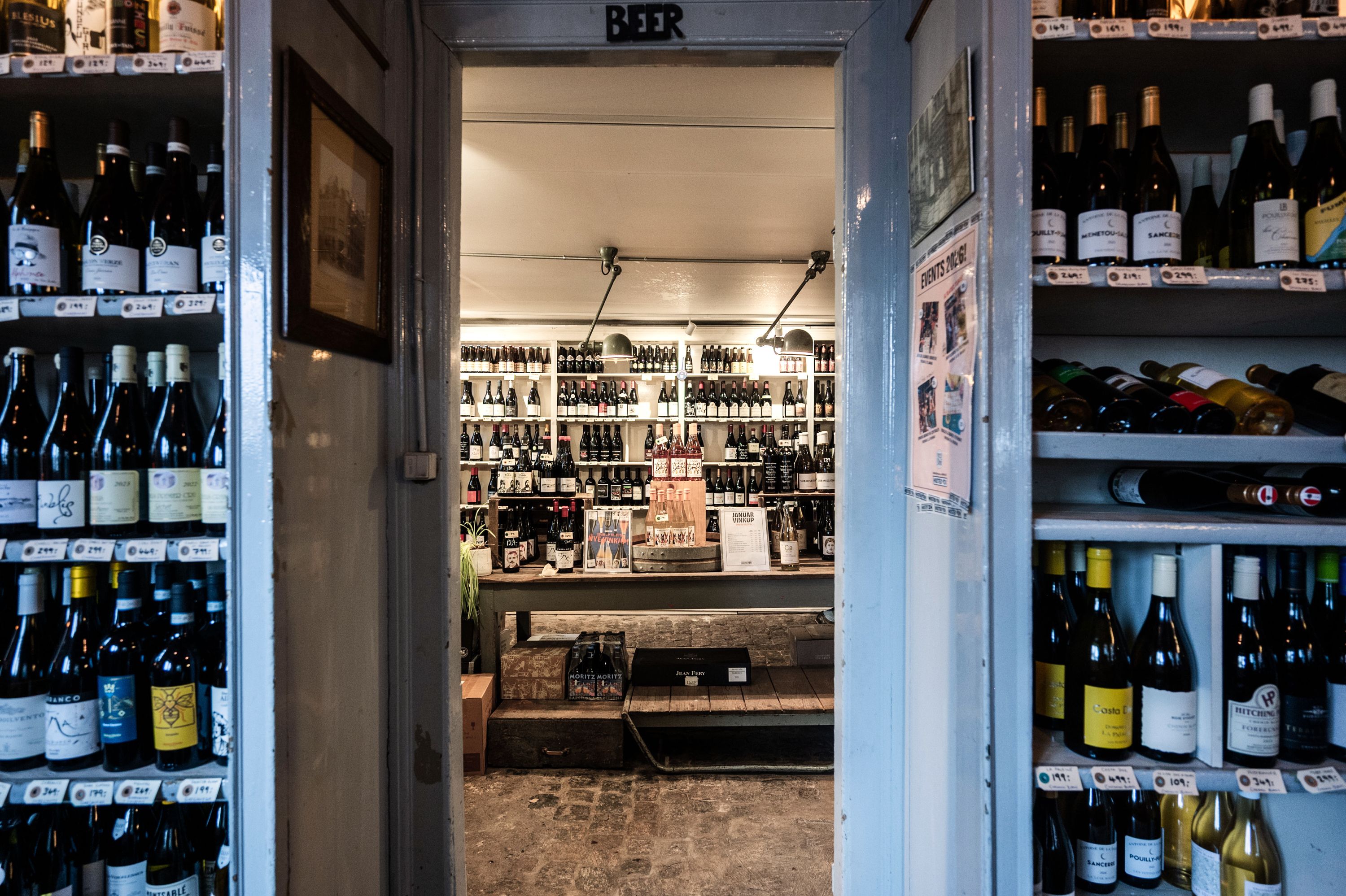 Store butikslokaler med lager og køkken samt hyggelige vin- og ølreoler, oplyst af lyse lyskatler, og åbne døre med udstilling af krystalflasker og etiketter.