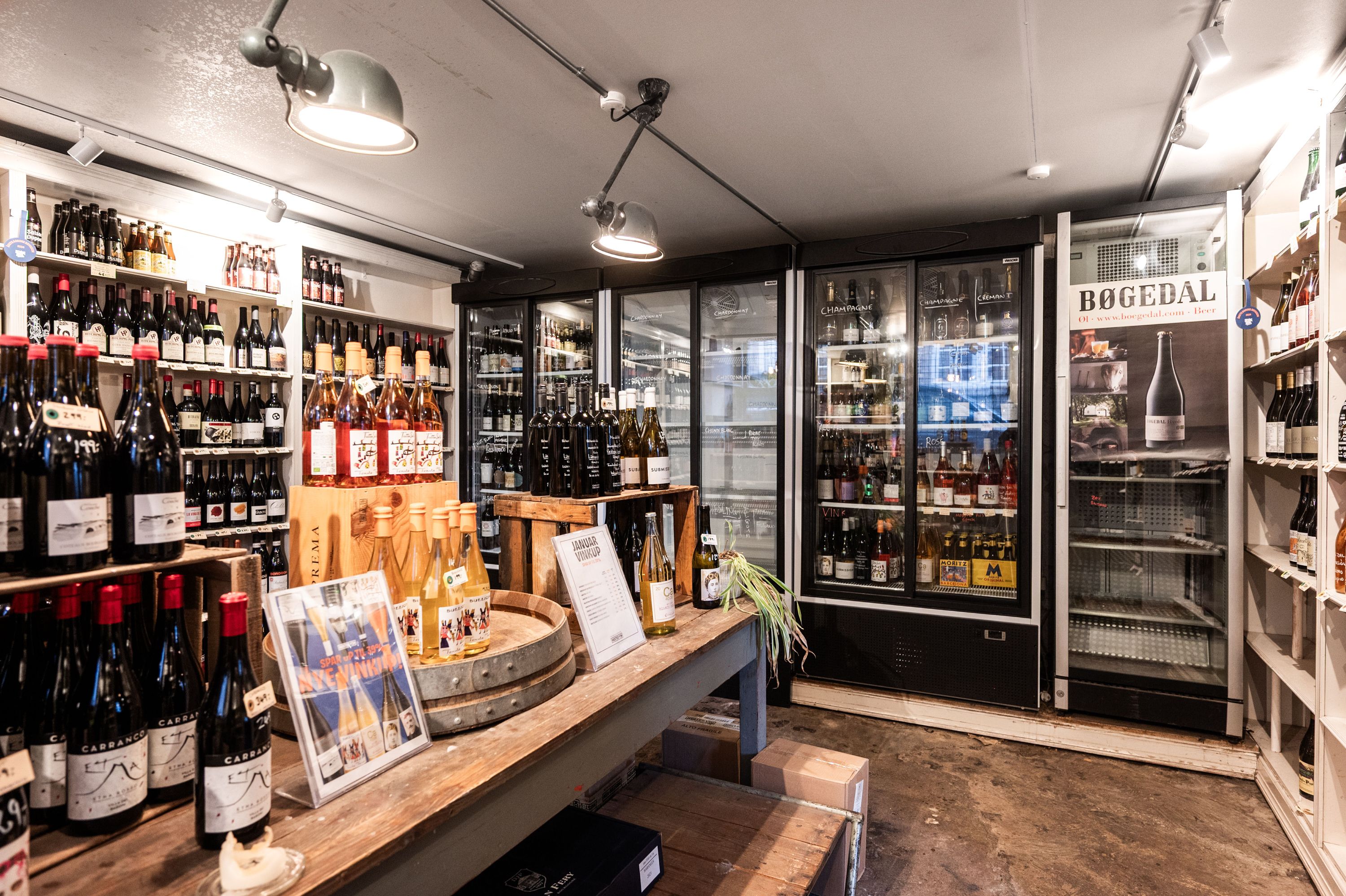 Butik med mange vinflasker i køleskabe og hylder og et stort træbord med en række vinflasker og nogle papæske.