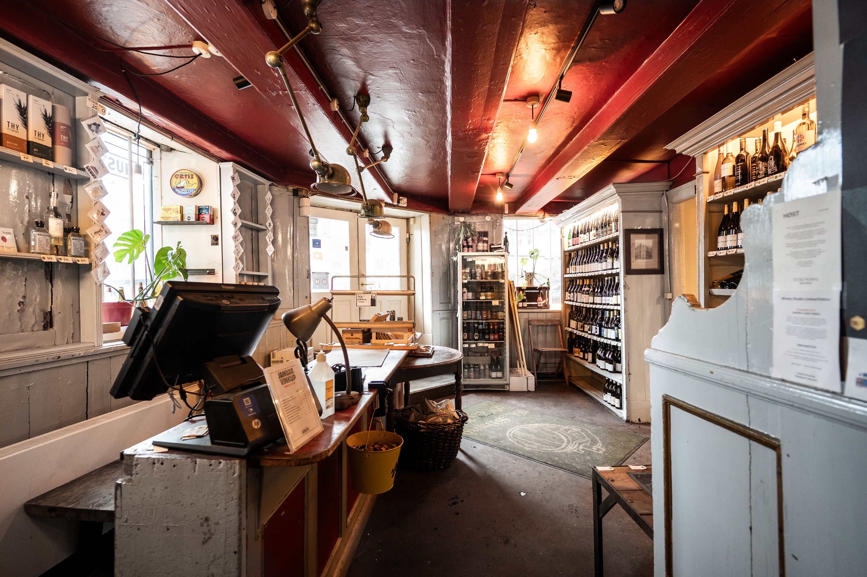 Indenfor en hyggeligt indrettet butik er der hylder med etiketter og glasflasker, en computer og en kasseapparat samt et gult spildtøj og en lampe