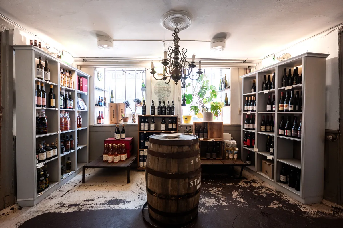 Vinbutik med mange etiketterede vinflasker på hylder og et stort tøndebord med hvid skive. Lys lysekroner hængende fra loftet.