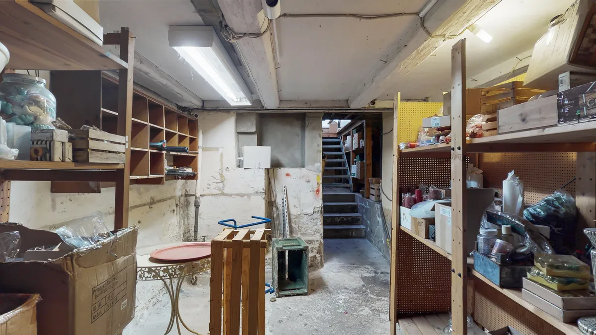 Et lagerrum med store hylder, bokse, trækasser og diverse genstande, inklusiv en glaskrukke og en klapvogn, lyst op af et hængende lys.