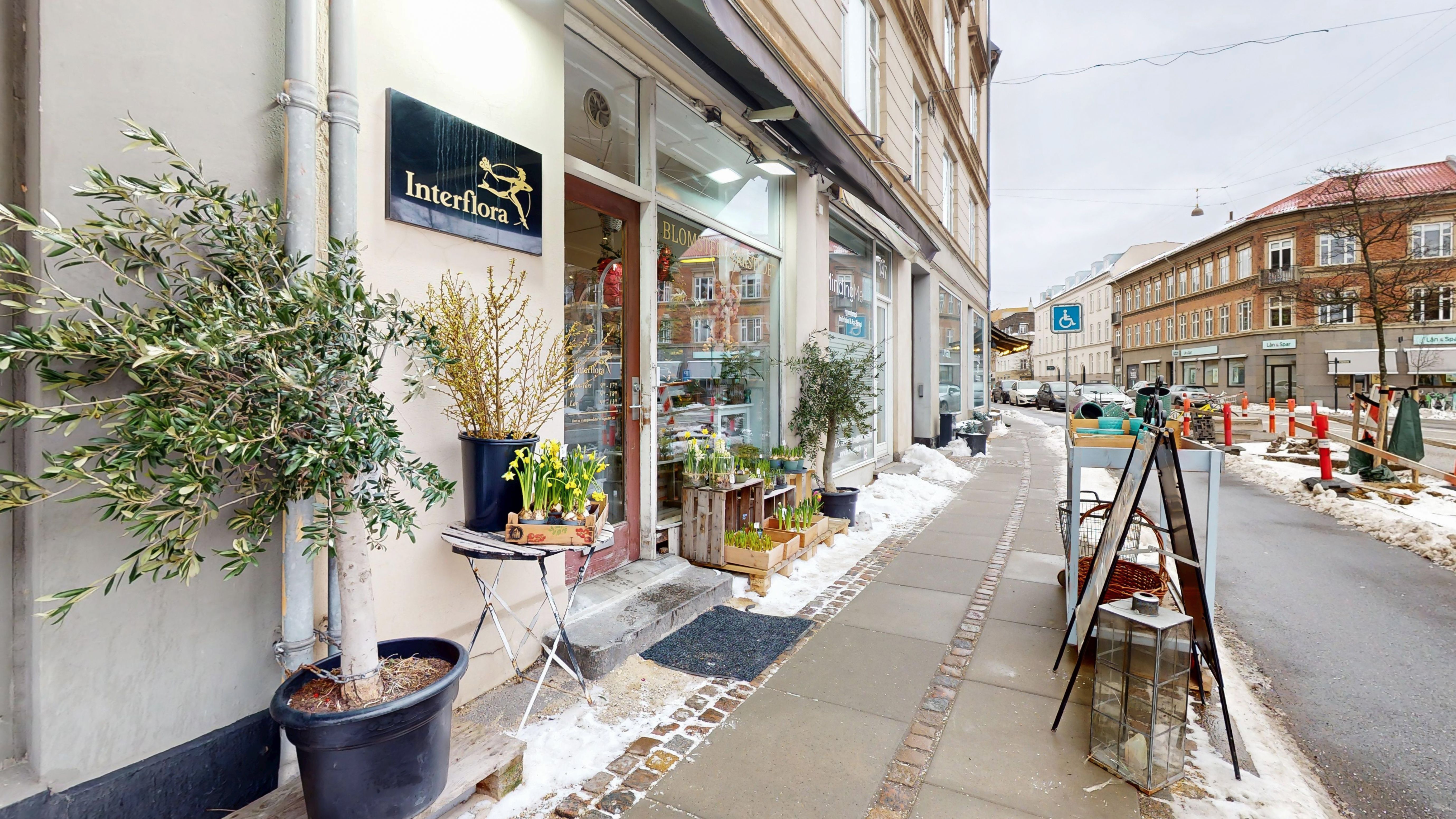 Butiksgade med Interflora-butik, blomsterpotter og sne på fortovet. Handicaptegn, kørestolsvenlig parkering og en række bygninger i baggrunden.