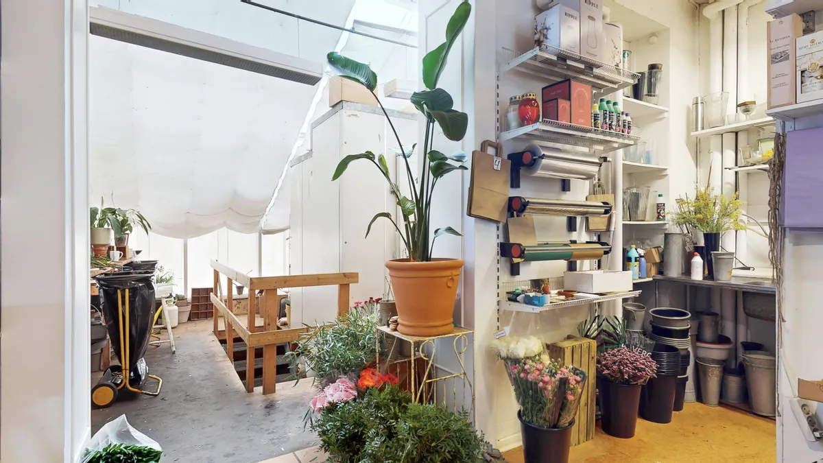 Indkøbsvogn og flere blomsterpotter i en veludstyret butik. På højre side er der en plante og en væg med hylder. Adgangsdør og trappe er i baggrunden.