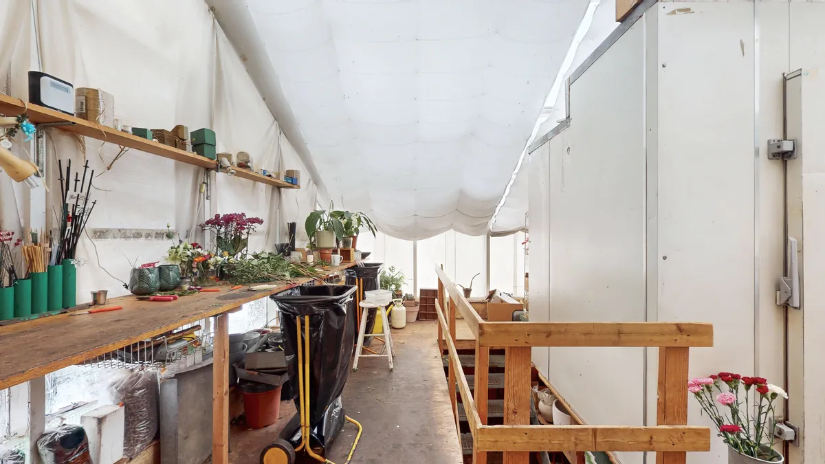 Indre af et velbeliggende erhvervslejemål med trægulve, højloft, lyseruder og blomster. Lejemålet indeholder værkstedsborde, potteplanter og støvsuger.