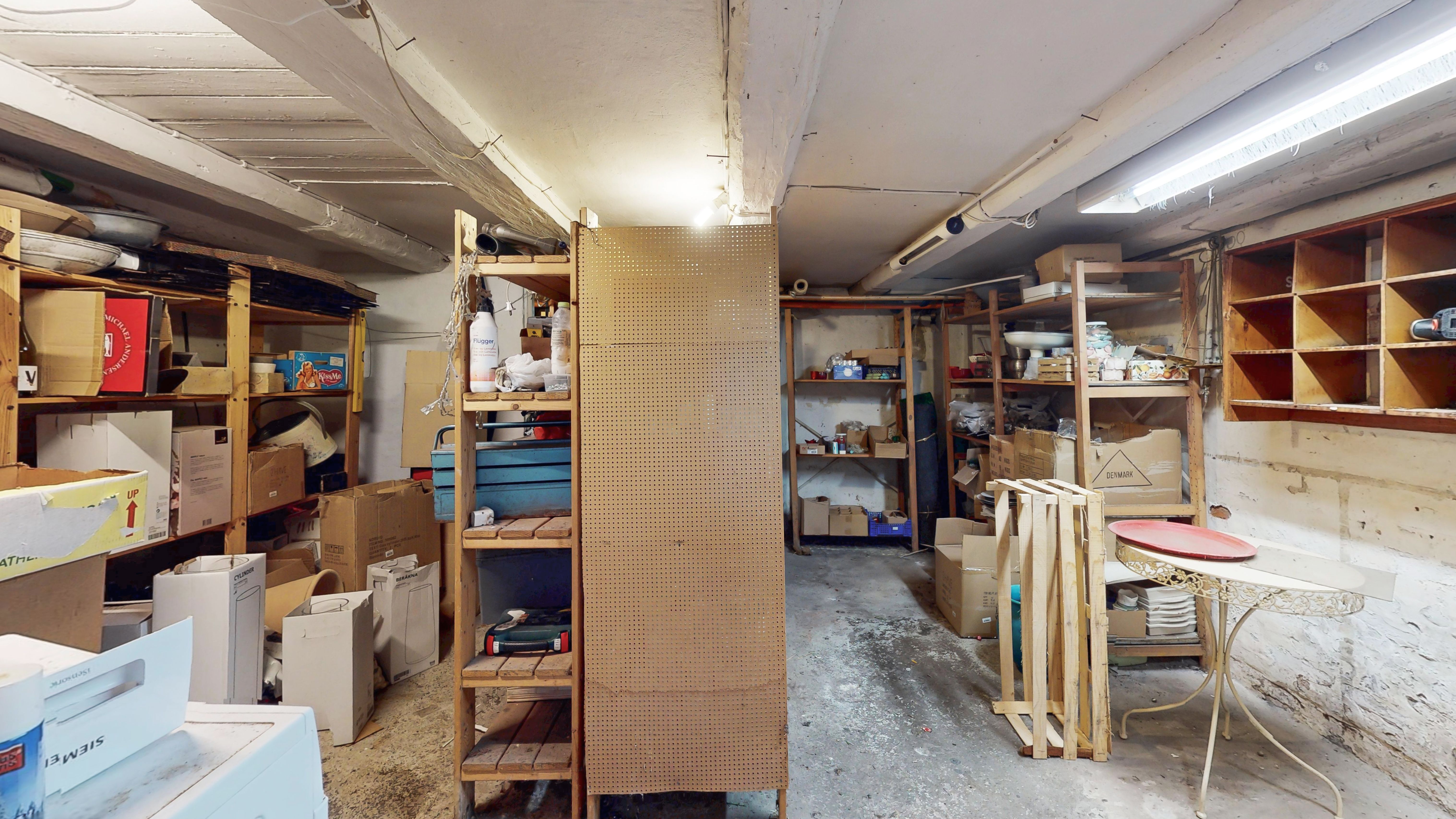 En lagerrum med hylder, kasser, værktøj og diverse ting. Der er en høj hældningsvæg og en lysearmatur på loftet.