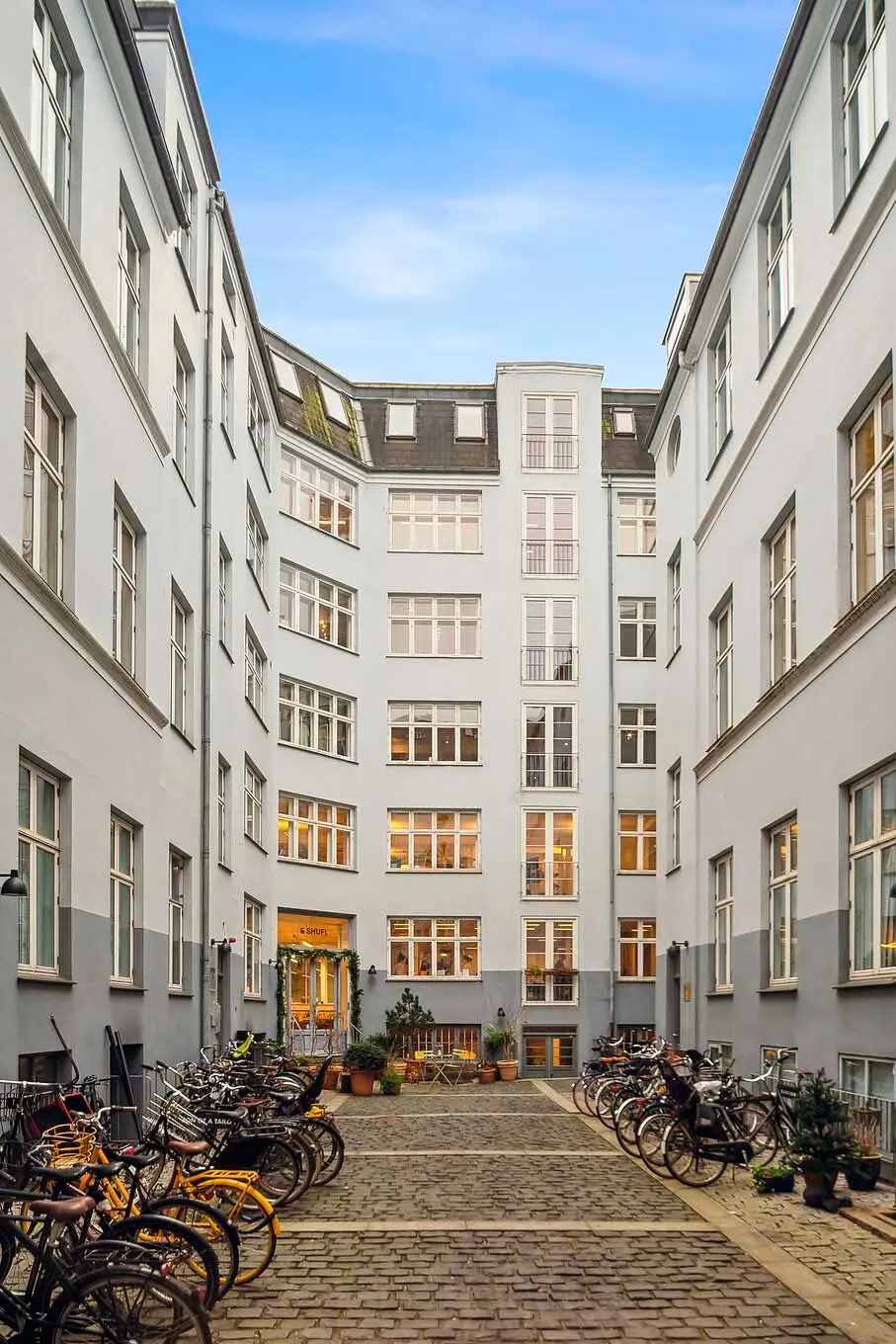 Bygning med flere etager og lyse vinduer i hjertet af København. Adgangsvej med gul gulvbelægning og parkerede cykler. Entrédør med skilt og grønne plantager i forgrunden