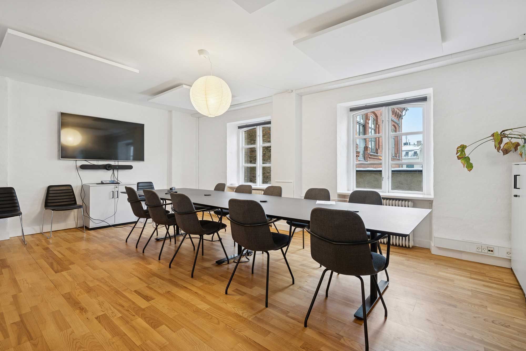 En kontormødelokale med en lang trægulv, moderne møbler og et stort fjernsyn på væggen. Et rundt lysekrogn hænger fra loftet, og der er vinduer med udsigt til bygninger.