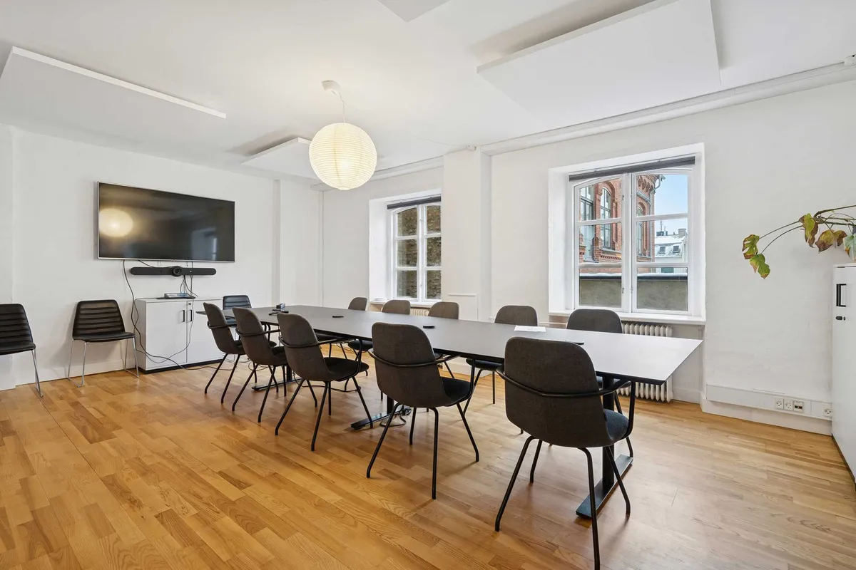 En kontormødelokale med en lang trægulv, moderne møbler og et stort fjernsyn på væggen. Et rundt lysekrogn hænger fra loftet, og der er vinduer med udsigt til bygninger.