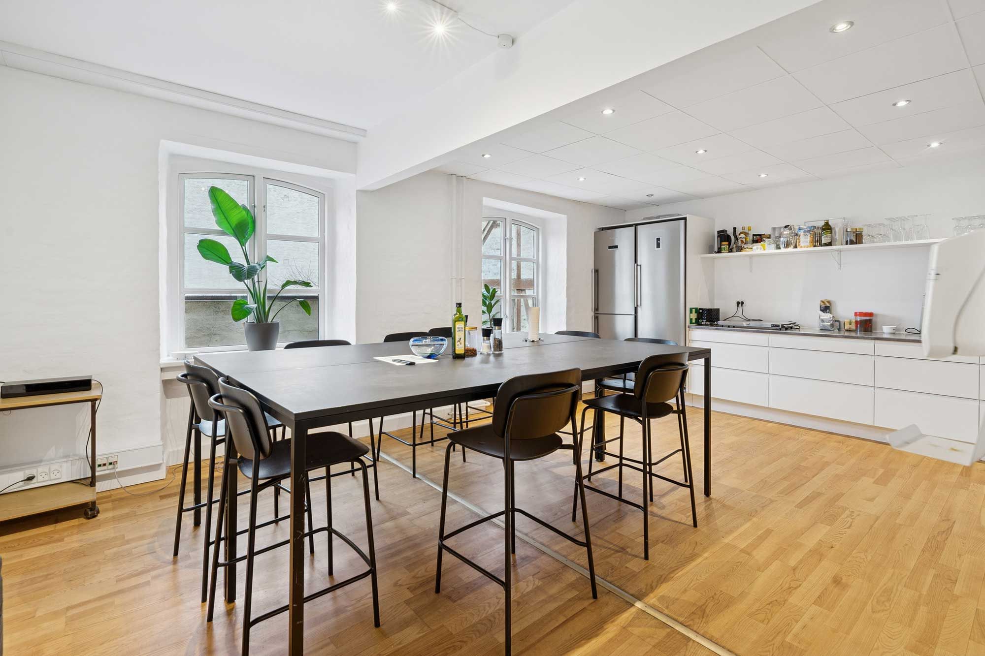 Kontor med spisebord og stole, hvidt køkken med indbygget køleskab og højt vinduesglas og et spisestuebord med plante og en flaske vin