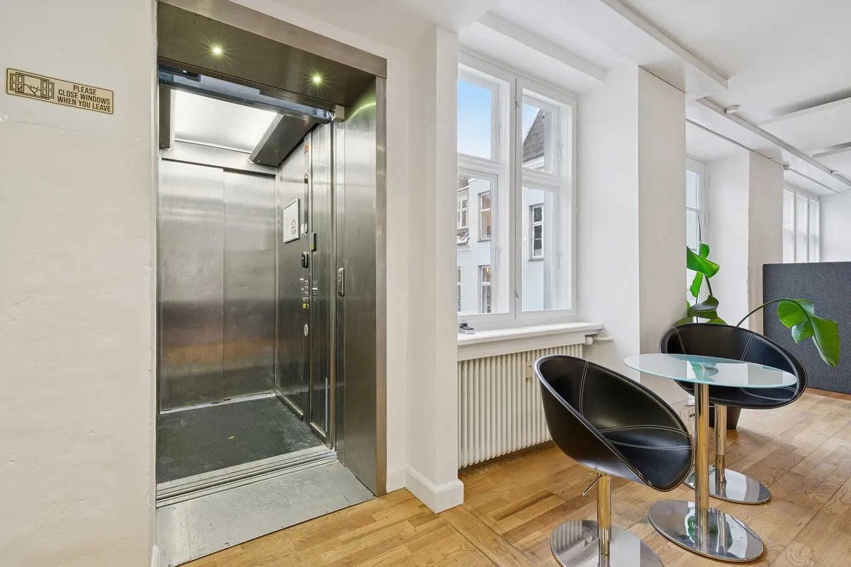 Kontor med elevator, lyse lokaler, to moderne læderstole og glasbord ved vinduet med udsigt til gaden og bygninger