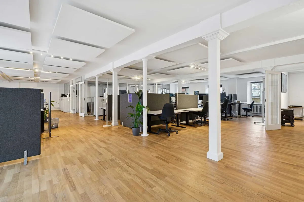 Kontor med højt loft og lyse gulve med søjler, moderne møbler og plante, sammen med et åbent rum og en dør