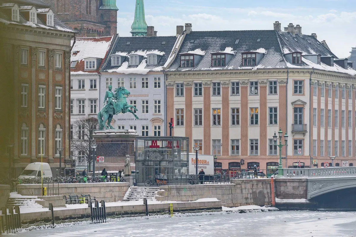 Bygninger med flere etager og statuer på kanalen i København. En vandkøretøj krydser kanalen, og et højt tårn er synligt i baggrunden.