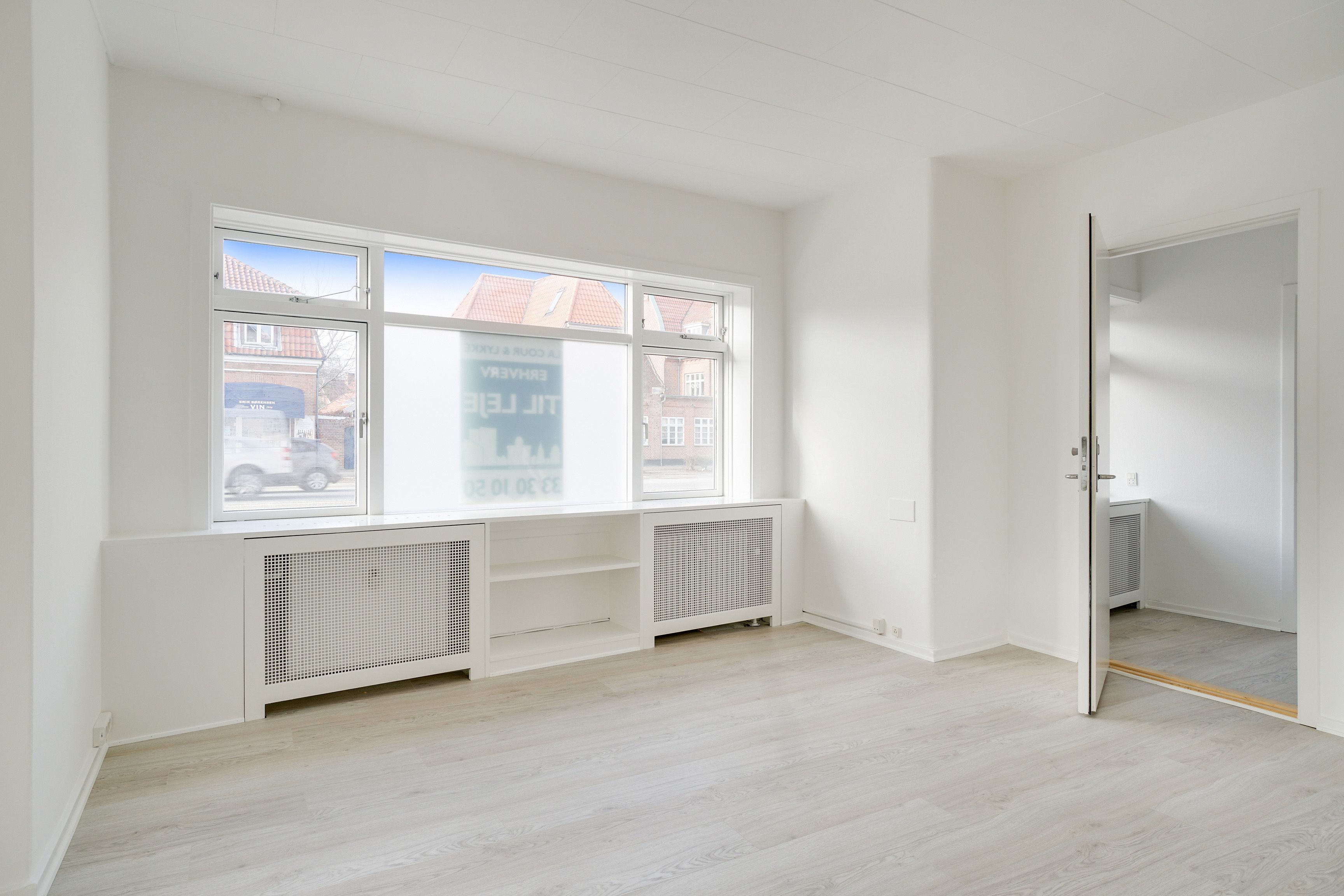 Lejlighed med lyse gulve og hvide vægge med vinduer og radiatorer. Et åbent dør og en hvid gulv i næste værelse. Og en reklame med "LET JETT" tekst på vinduet.