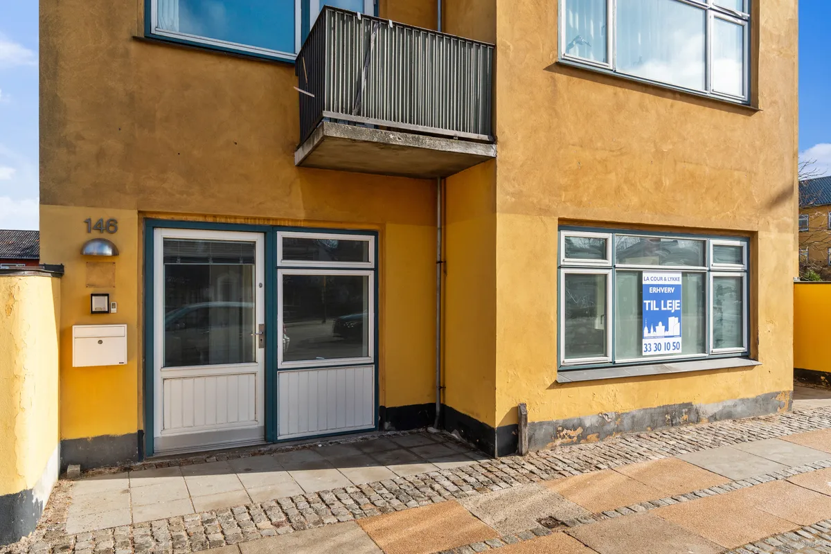 Kontor til leje på Bernstorffsvej 146 i Hellerup, med oplysninger om arealet og lejeprisen. Lysgul facade med vinduer og balkon, samt en dør med mailkasse og nummerplade.