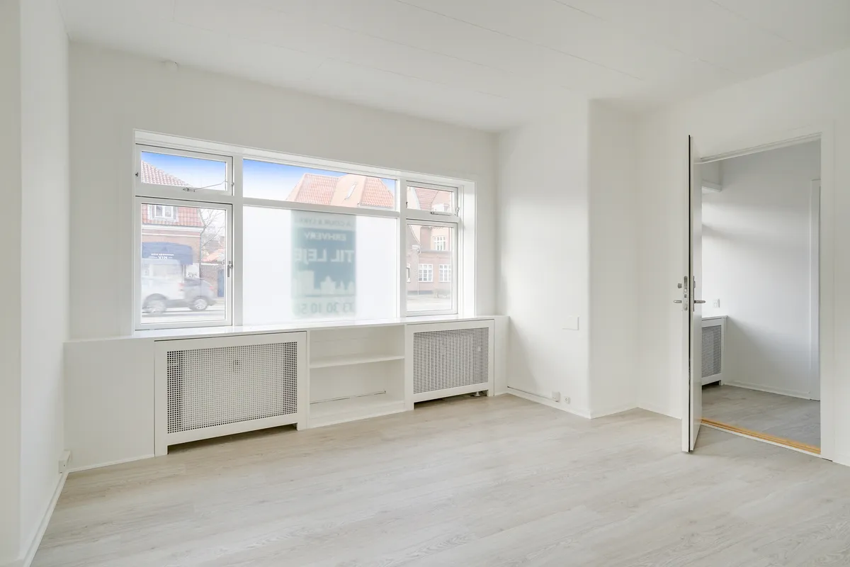 Et lejlighedsværelse med hvide vægge og gulv samt rektangulært vindue med glas. Der er radiatorer under vinduet og skrift på det ene af vinduetsruder.
